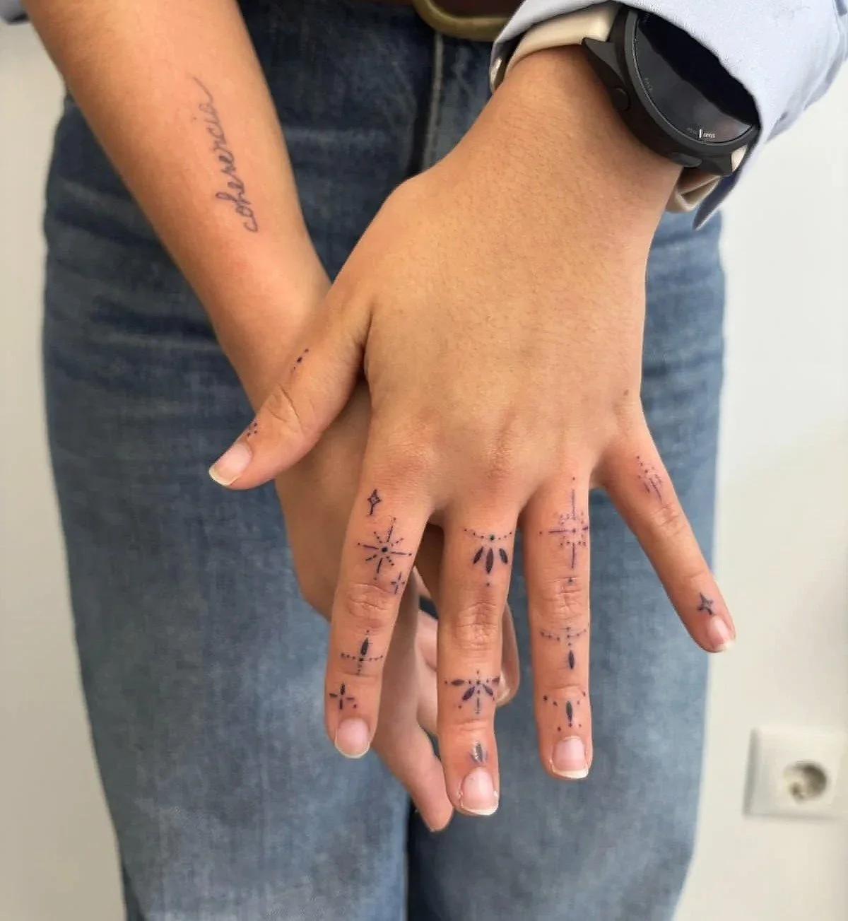 Persona con tatuajes en los dedos y en el brazo, usando un reloj negro y ropa informal, en un fondo de pared blanca.