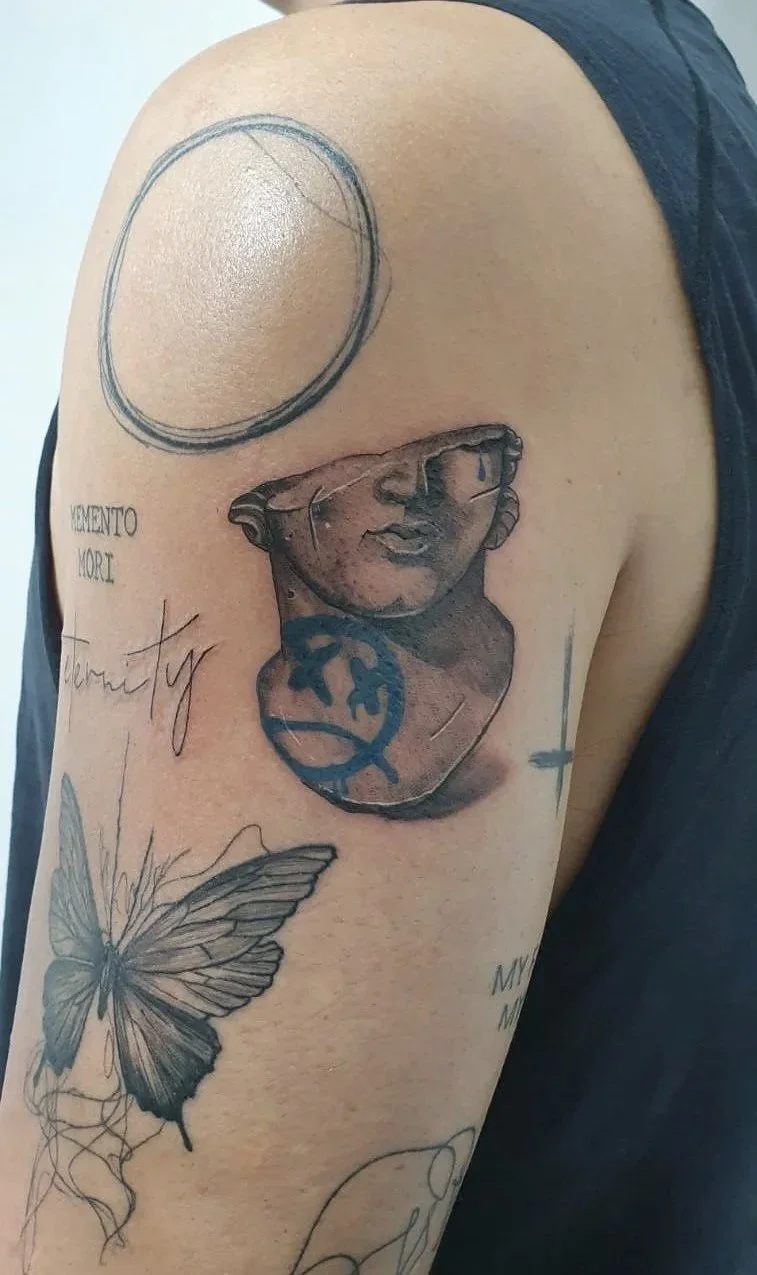 Tatuaje en el hombro de una mujer que incluye un busto de una figura clásica, una mariposa, palabras en diferentes estilos y algunos círculos.