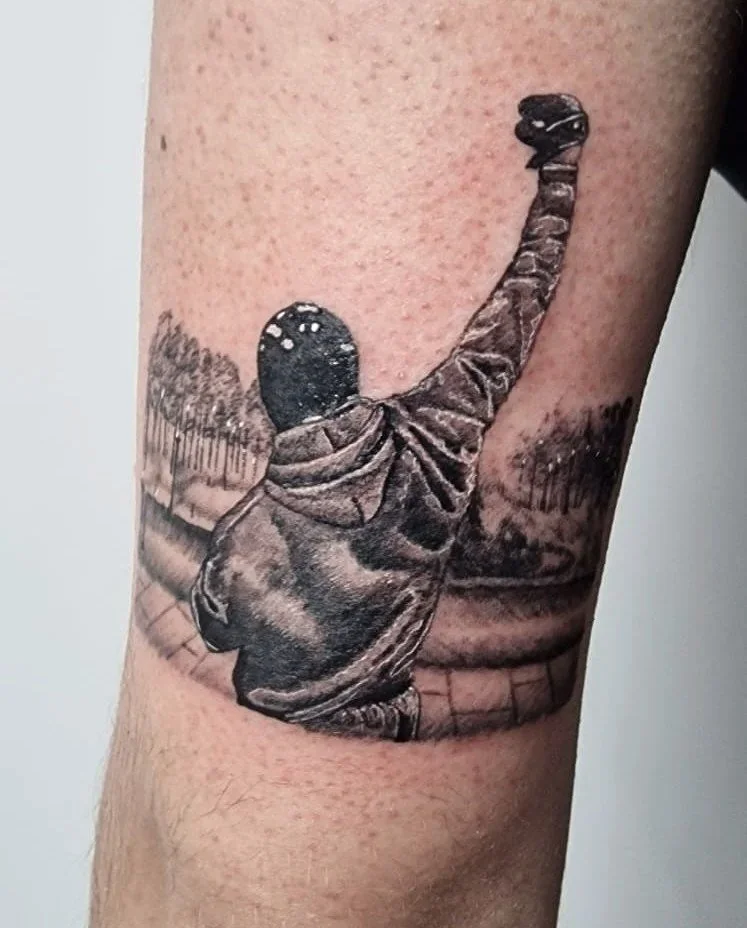 Tatuaje en tono de gris de una figura que parece un astronauta con el brazo levantado y un fondo de paisaje con árboles y un camino.