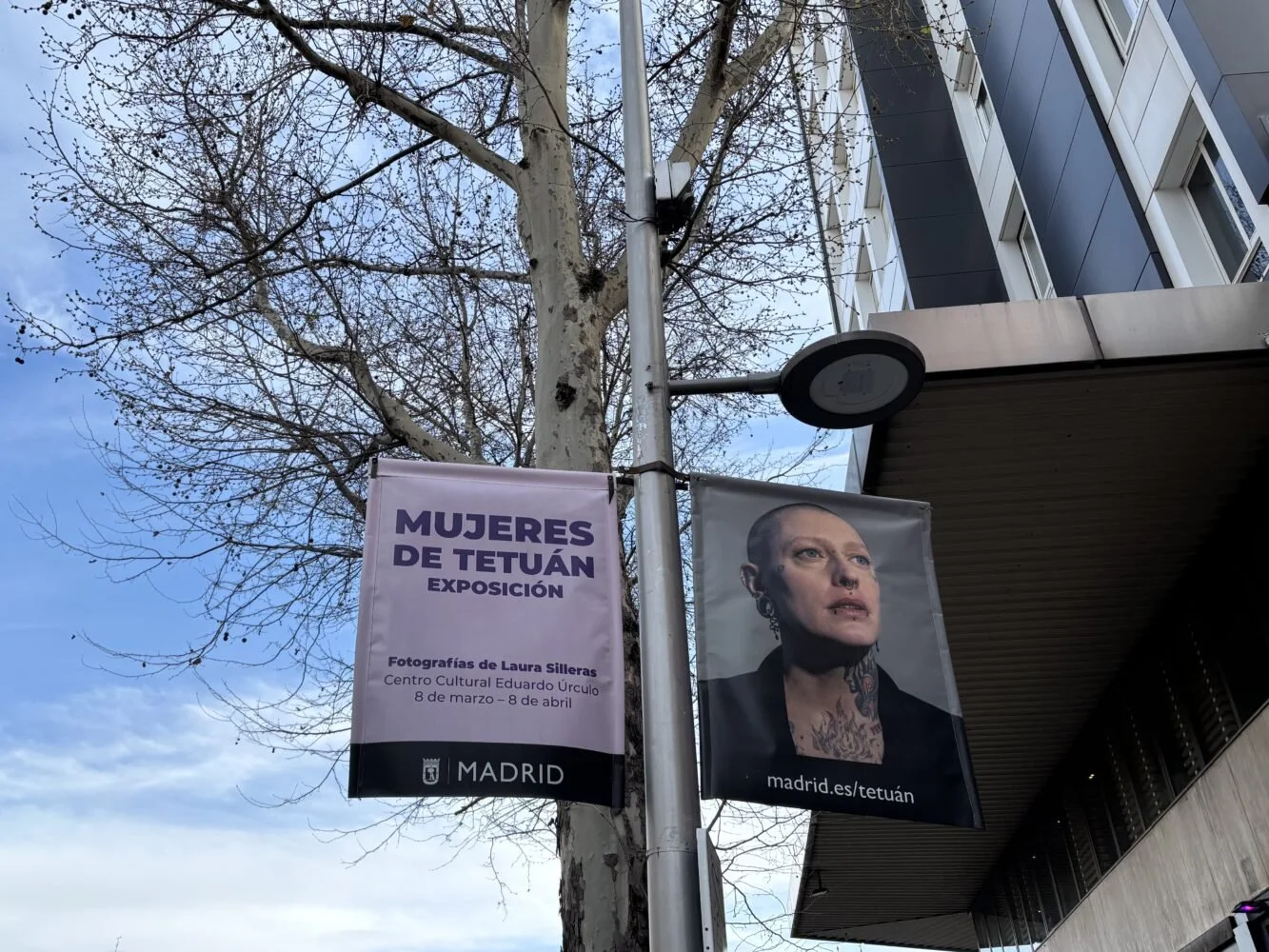 Carteles publicitarios para una exposición fotográfica llamada Mujeres de Tetuán en Madrid, con detalles de la exposición y una foto de una mujer con tatuajes en el cuello y cara.
