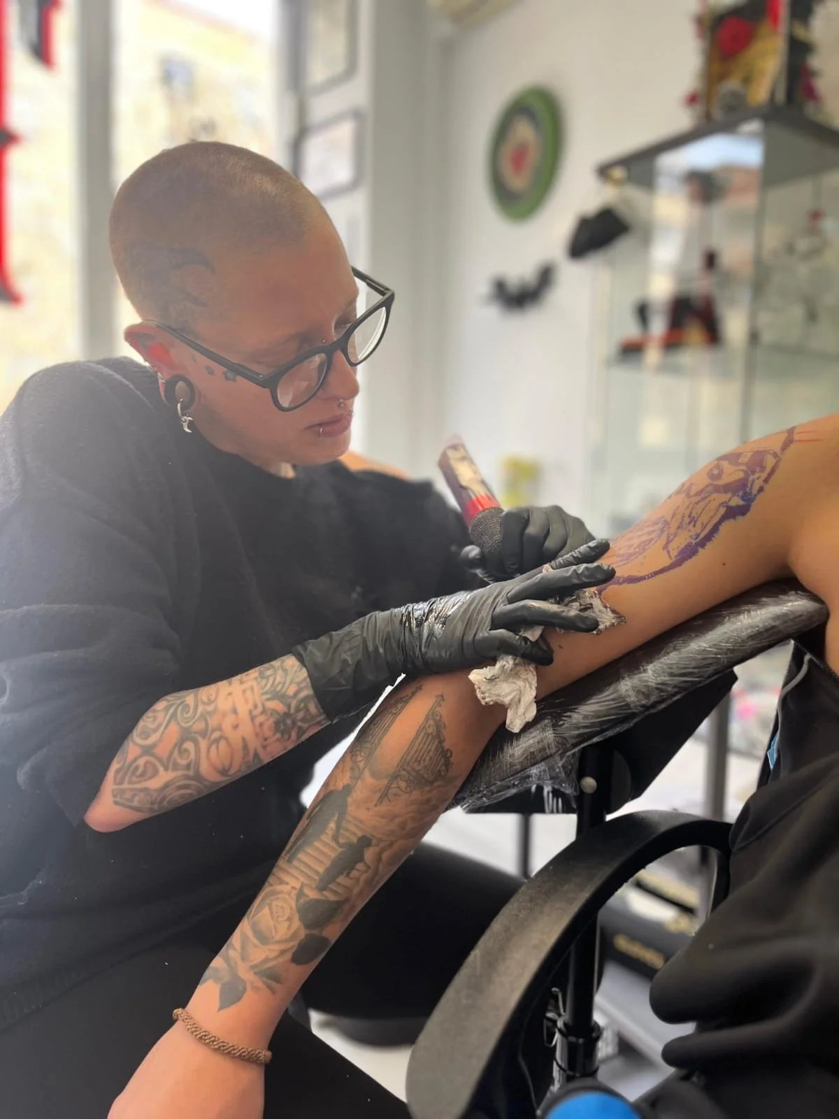 Una mujer con tatuajes en los brazos, gafas y piercings, tatúa a una persona en el brazo en un estudio de tatuajes.