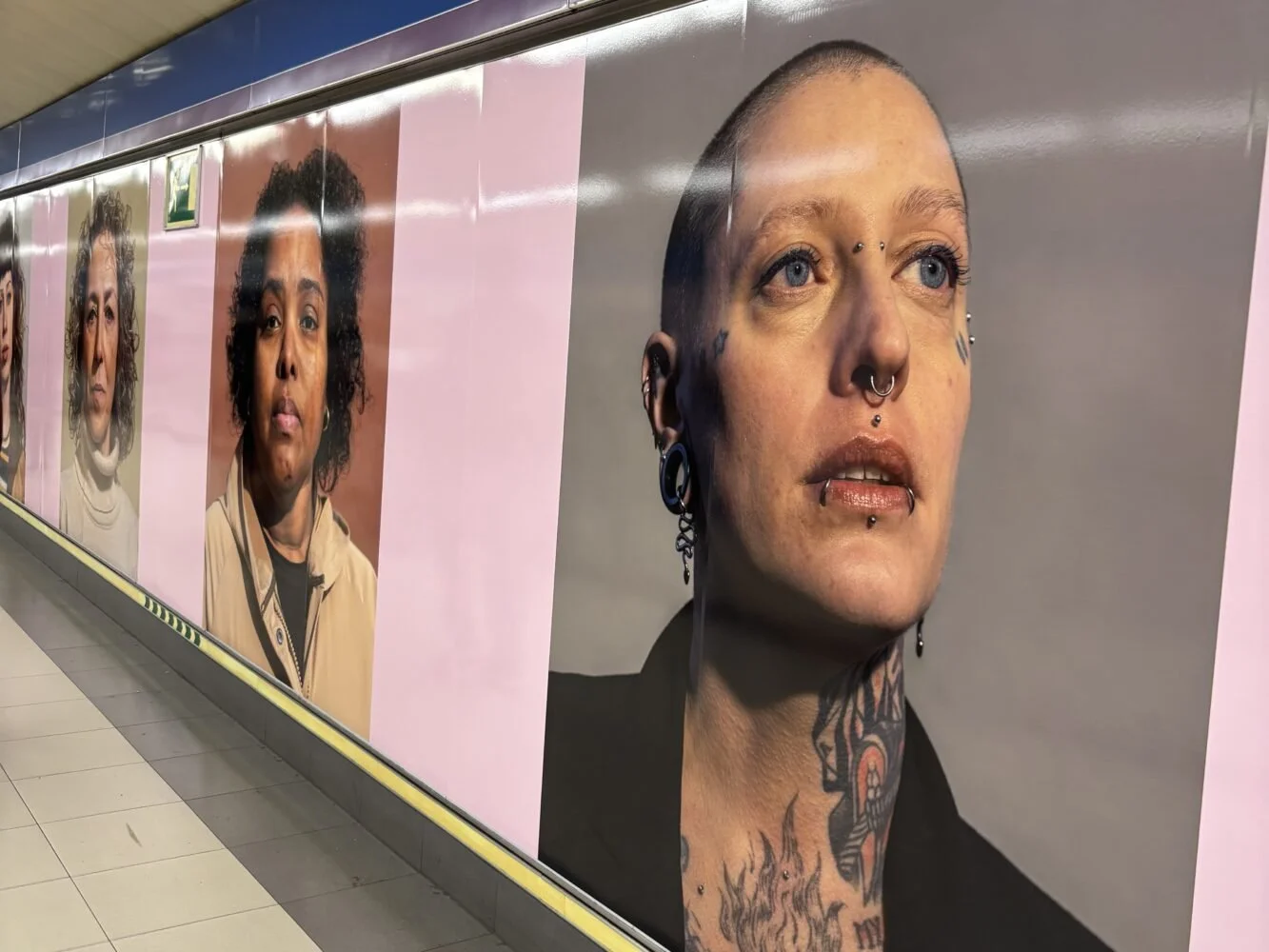 Imágenes de retratos de varias personas con diferentes estilos y expresiones, decoradas con piercings y tatuajes, en un mural público.