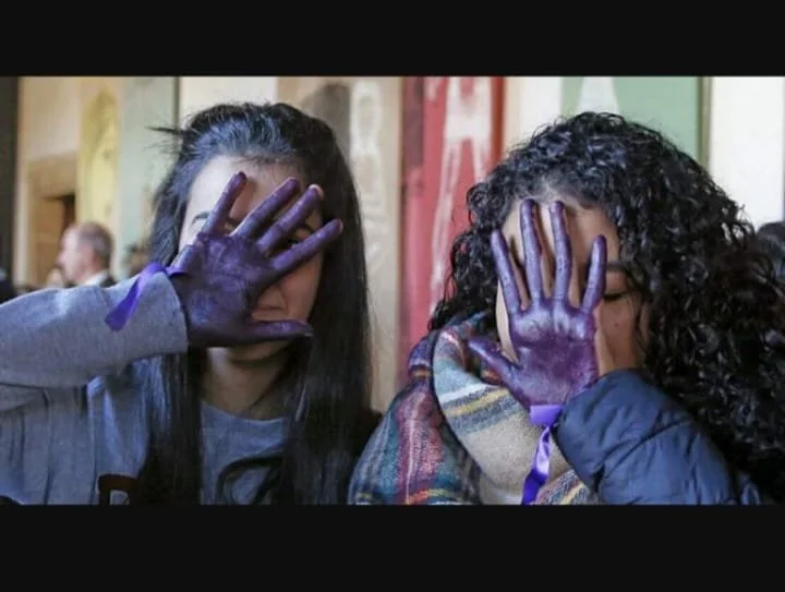 Dos jóvenes con las manos pintadas de morado cubriéndose los rostros en un ambiente interior.