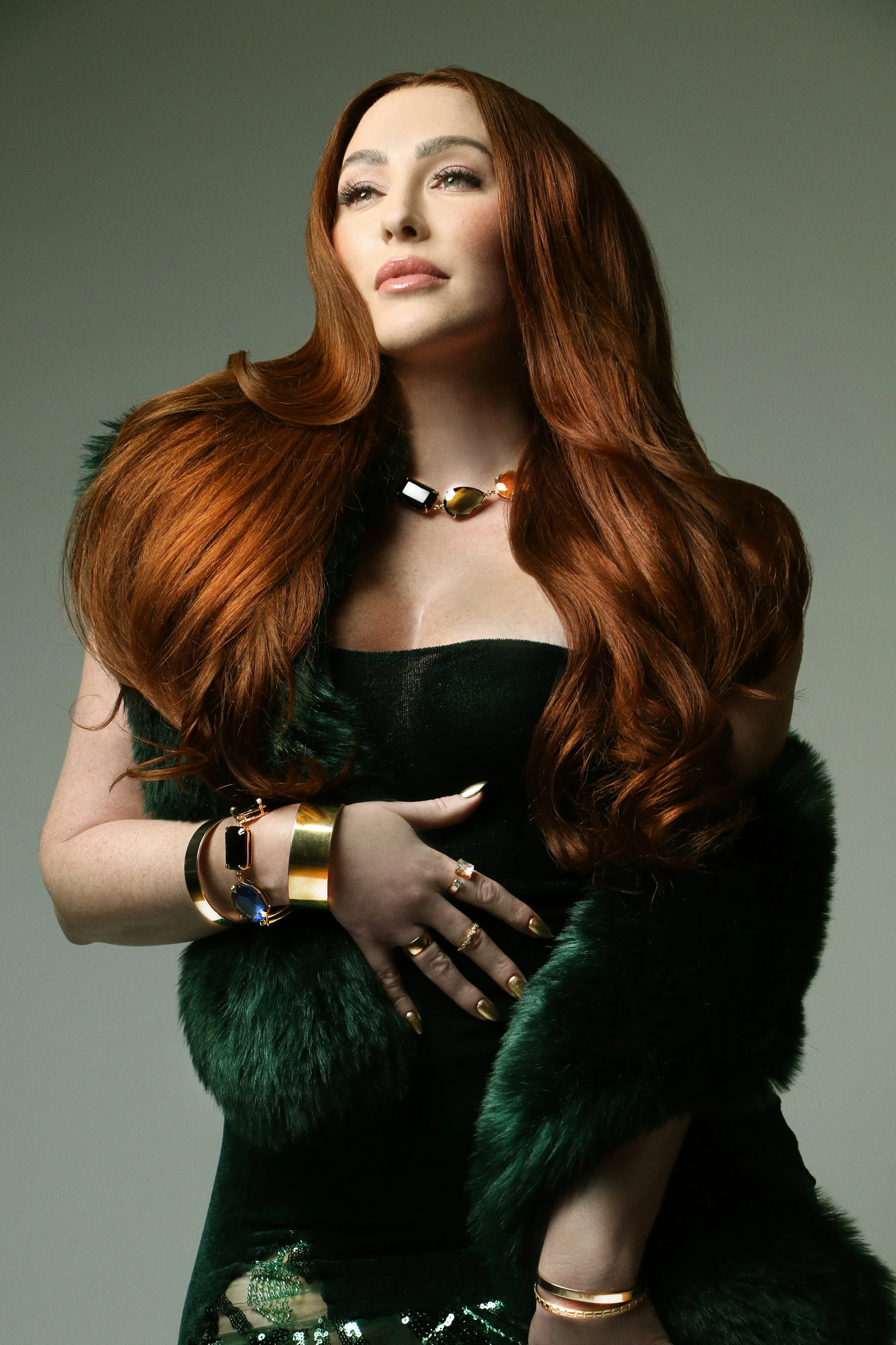 A woman with long, wavy red hair wearing a black strapless dress and green fur stole. She is adorned with gold jewelry including rings, bracelets, and a necklace, and has a confident expression.