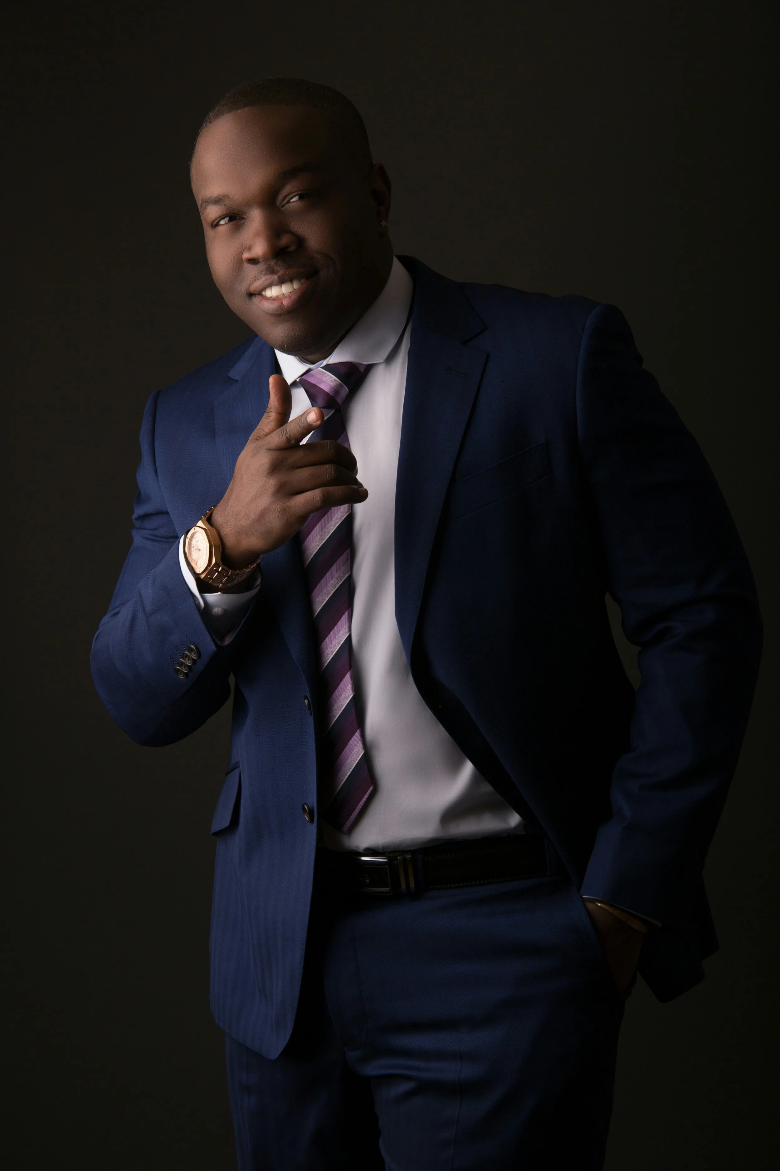 A man in a dark blue suit with a white shirt and a striped tie, posing confidently with his right hand near his chin and his left hand in his pocket, against a dark background.