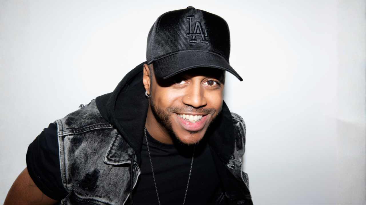 Bradley from S Club wearing a black Los Angeles cap, a black hoodie, and a denim vest, posing against a plain white background.