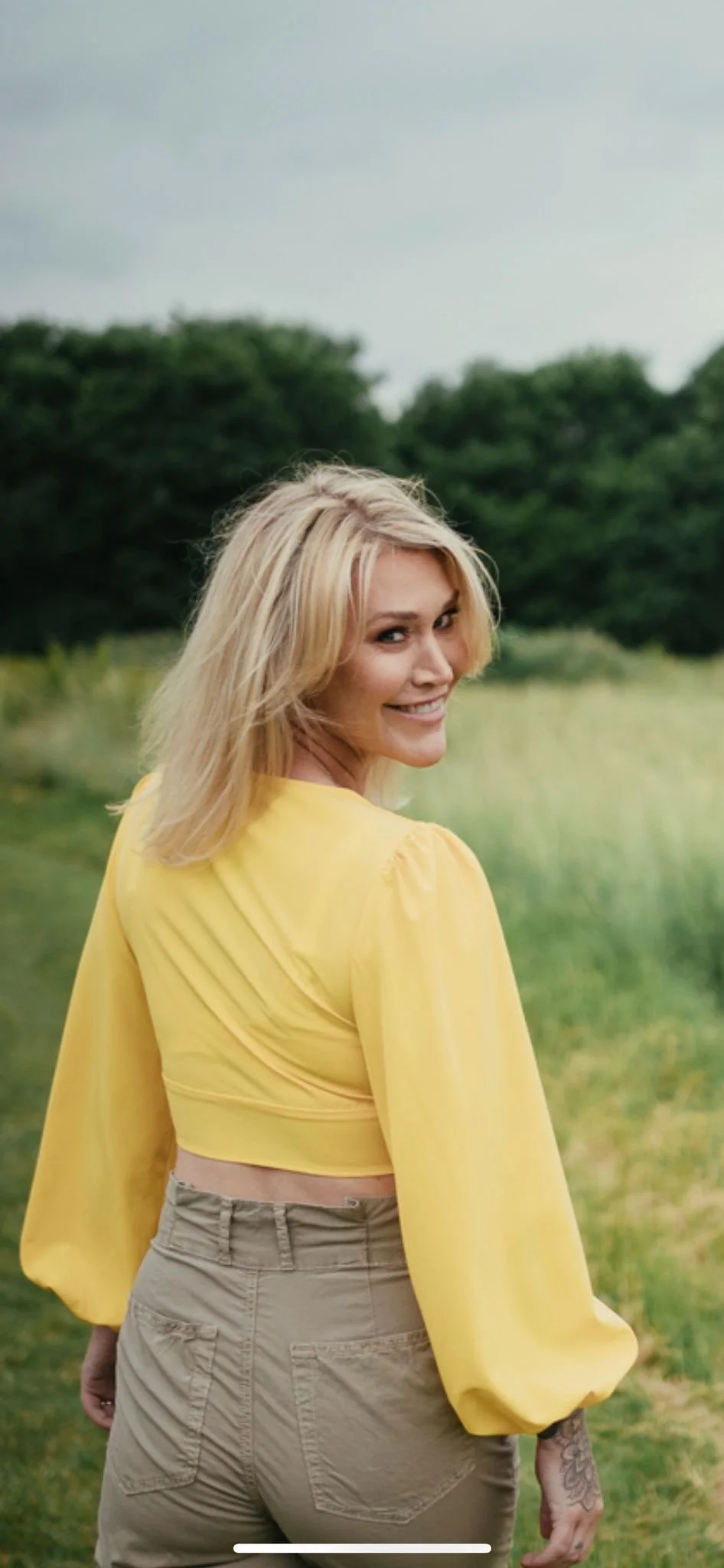 A woman with blonde hair wearing a yellow long-sleeve cropped top and khaki pants, smiling and looking back in a grassy field with trees in the background.