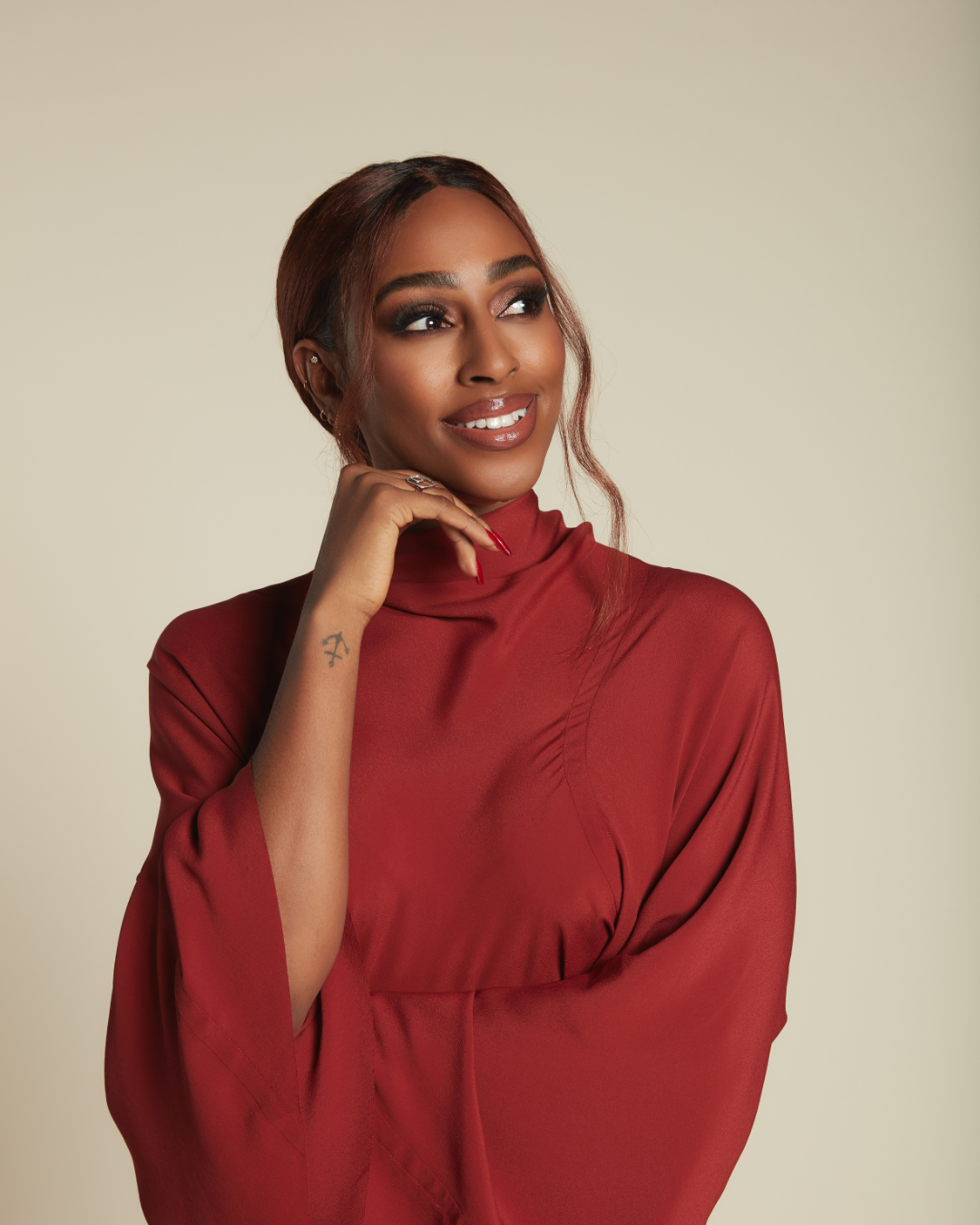 Alexandra Burke with brown hair styled in loose waves, wearing a red satin top with a turtleneck, smiling and looking to the side with her hand near her chin, standing against a light beige background.