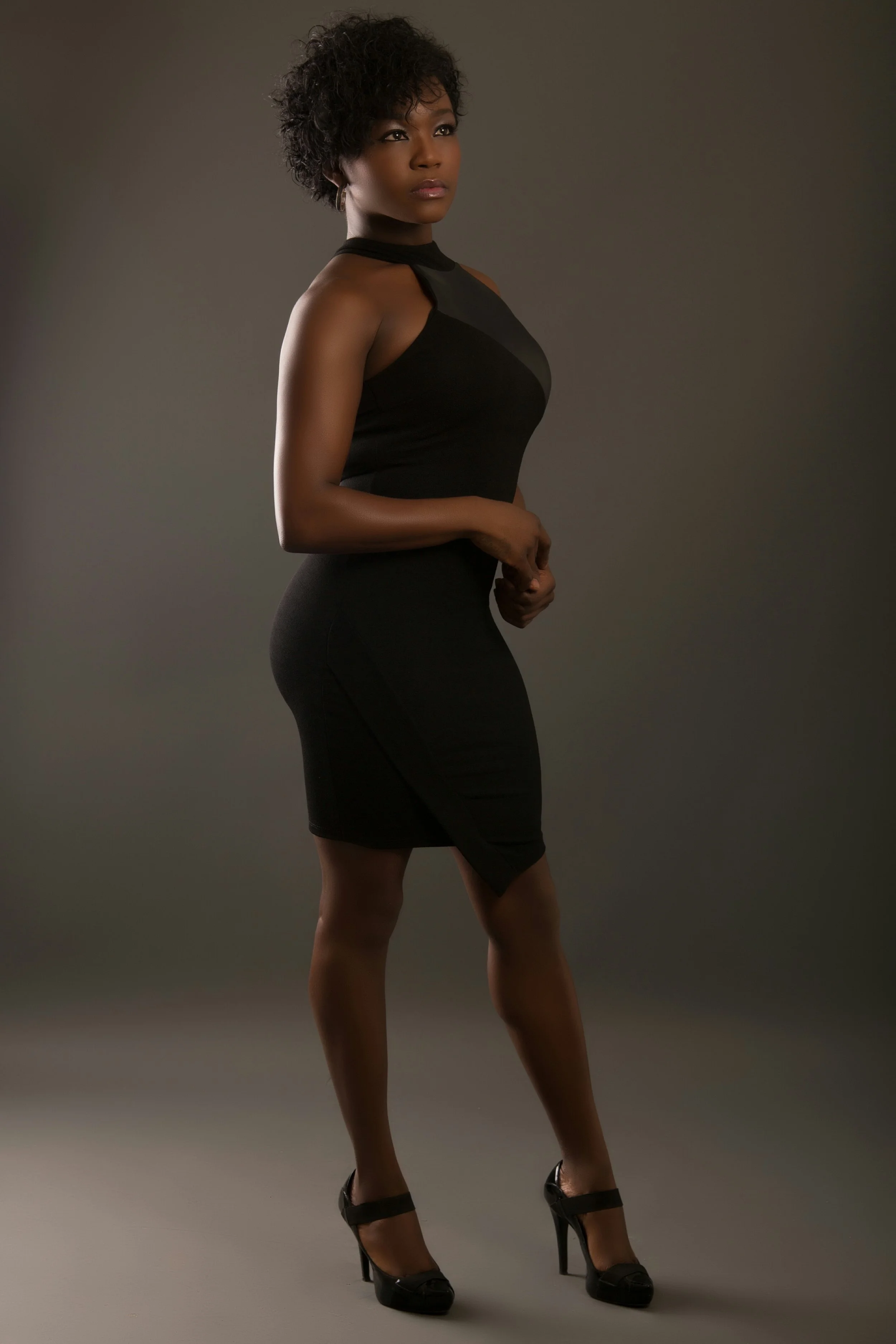 A woman wearing a fitted black dress and high heels, posed against a neutral background.