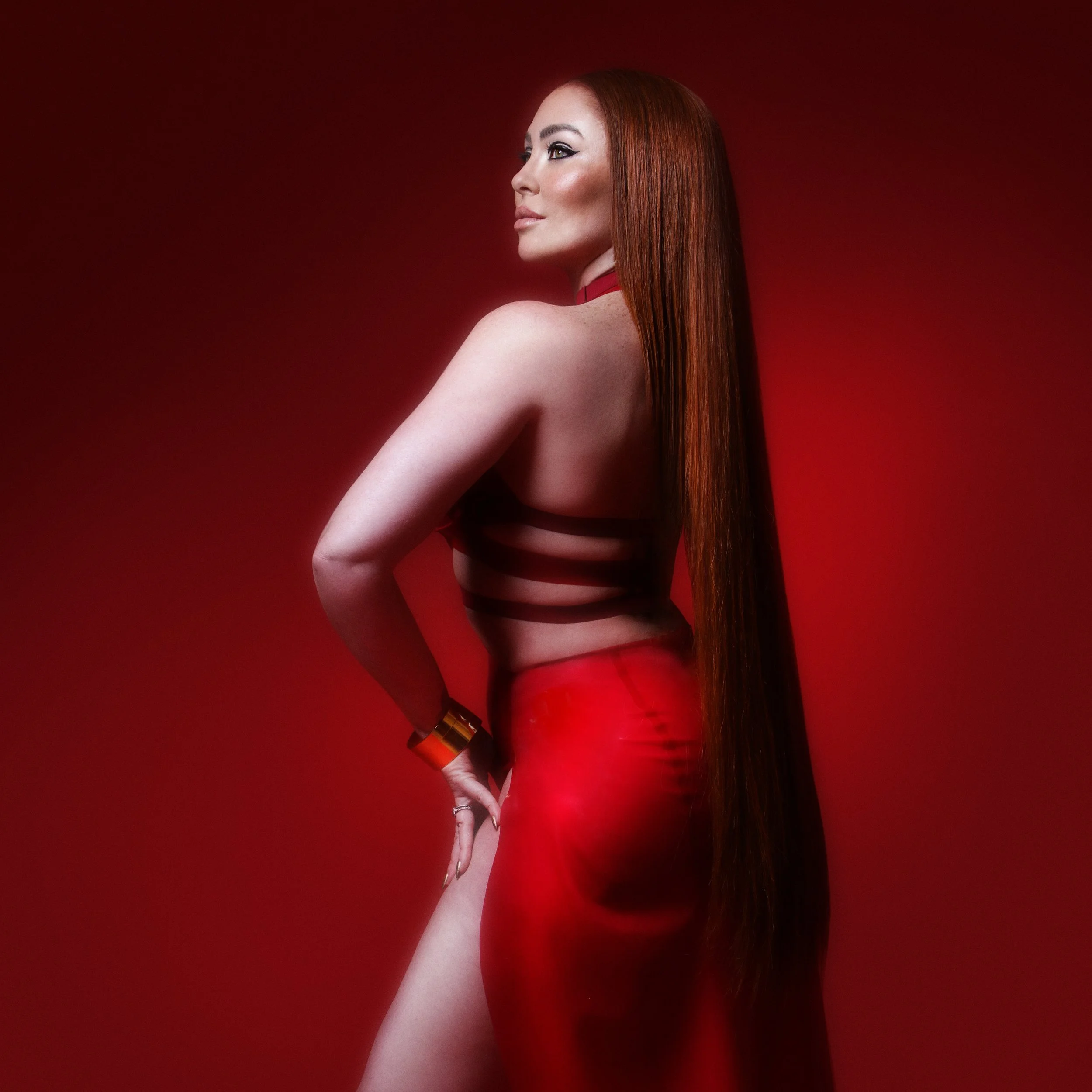 A woman with long red hair posing against a red background, wearing a black top and red skirt.