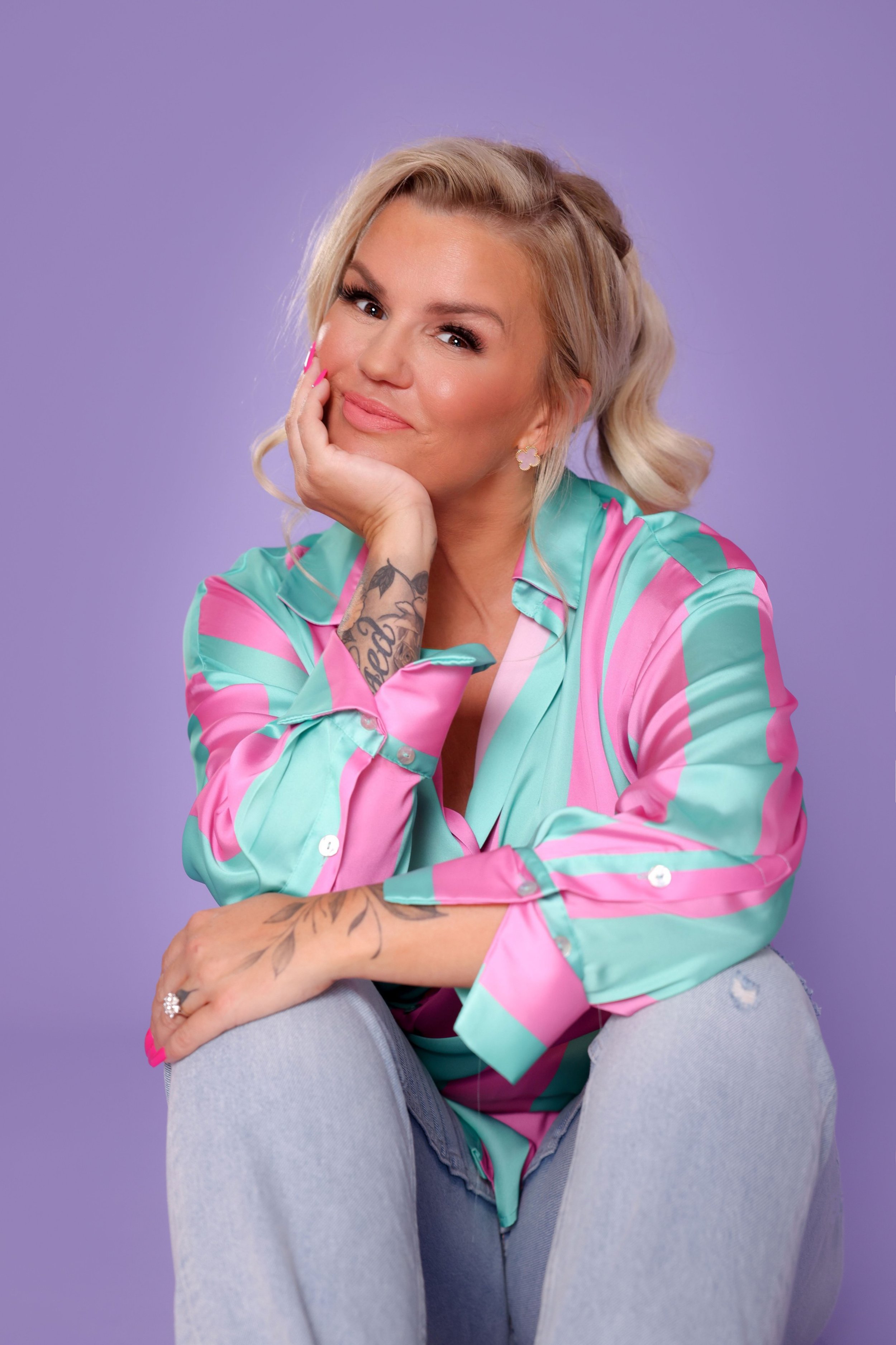 Kerry Katona wearing a light pastel-colored striped button-up shirt and light blue jeans, sitting against a purple background. She is resting her face in her right hand, with her elbow on her knee, and is smiling softly.