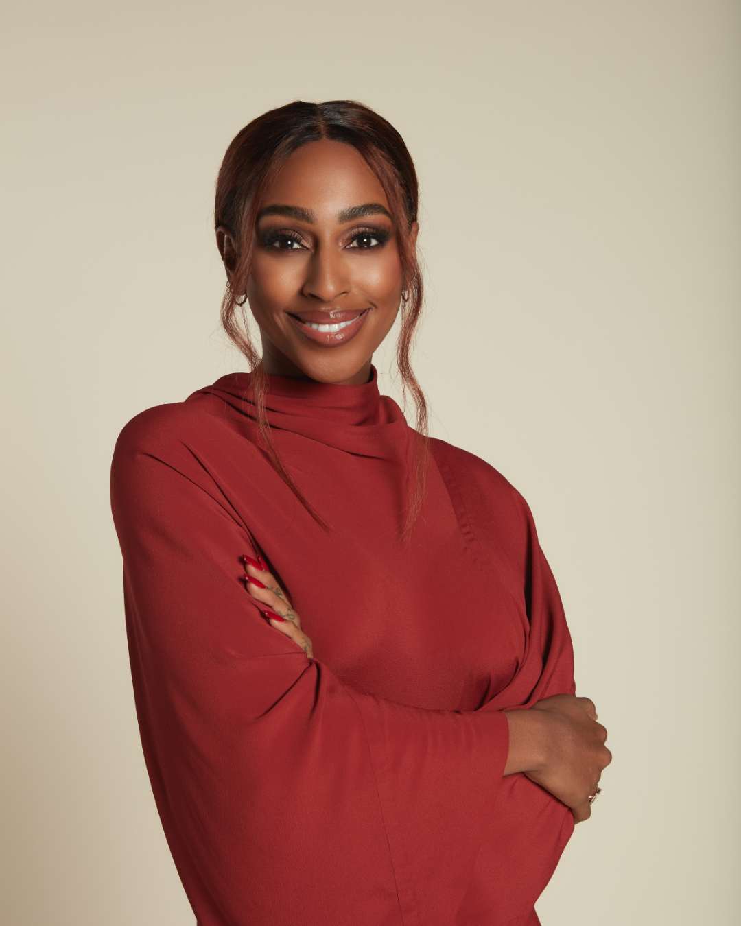 A woman with brown skin, wearing a red long-sleeved top, smiling and with arms crossed against a light-colored background.