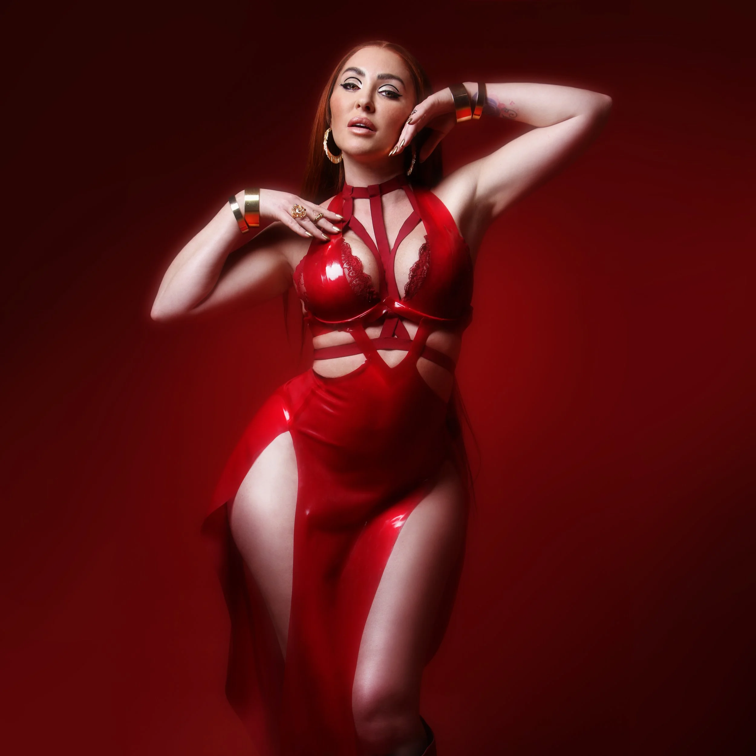 A woman in a revealing red lace and latex dress striking a pose against a red background.