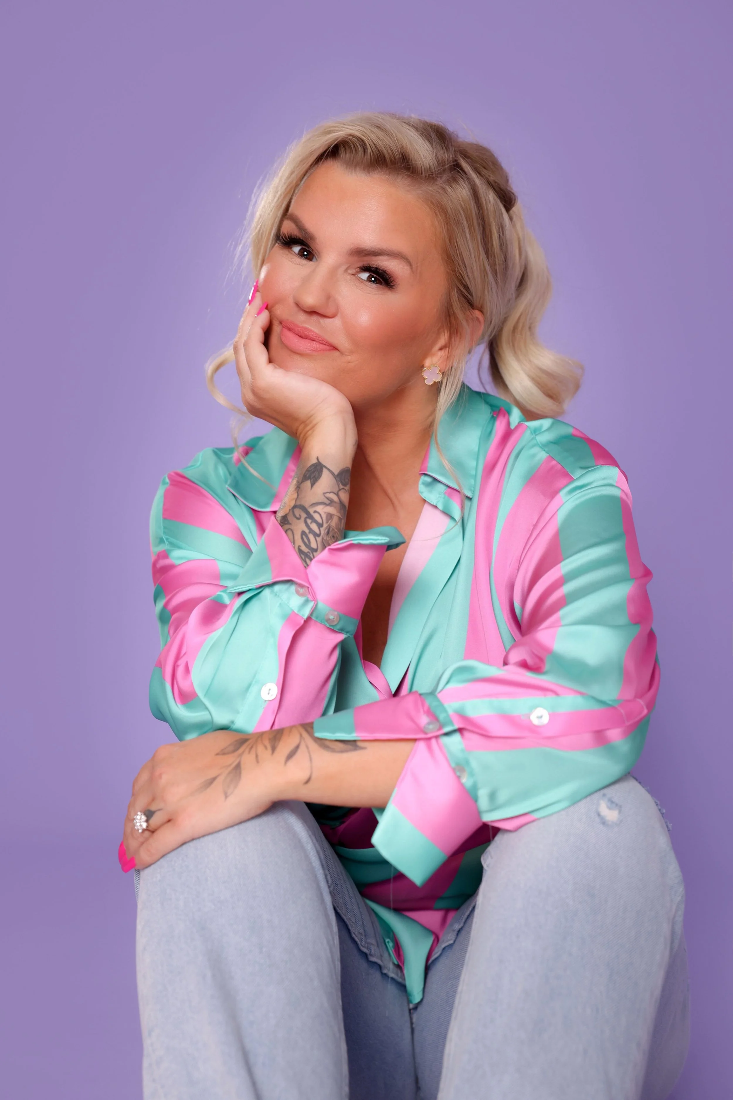 A woman with blonde hair, tattoos, and earrings, dressed in a colorful satin shirt and light jeans, sitting on the floor with a purple background.
