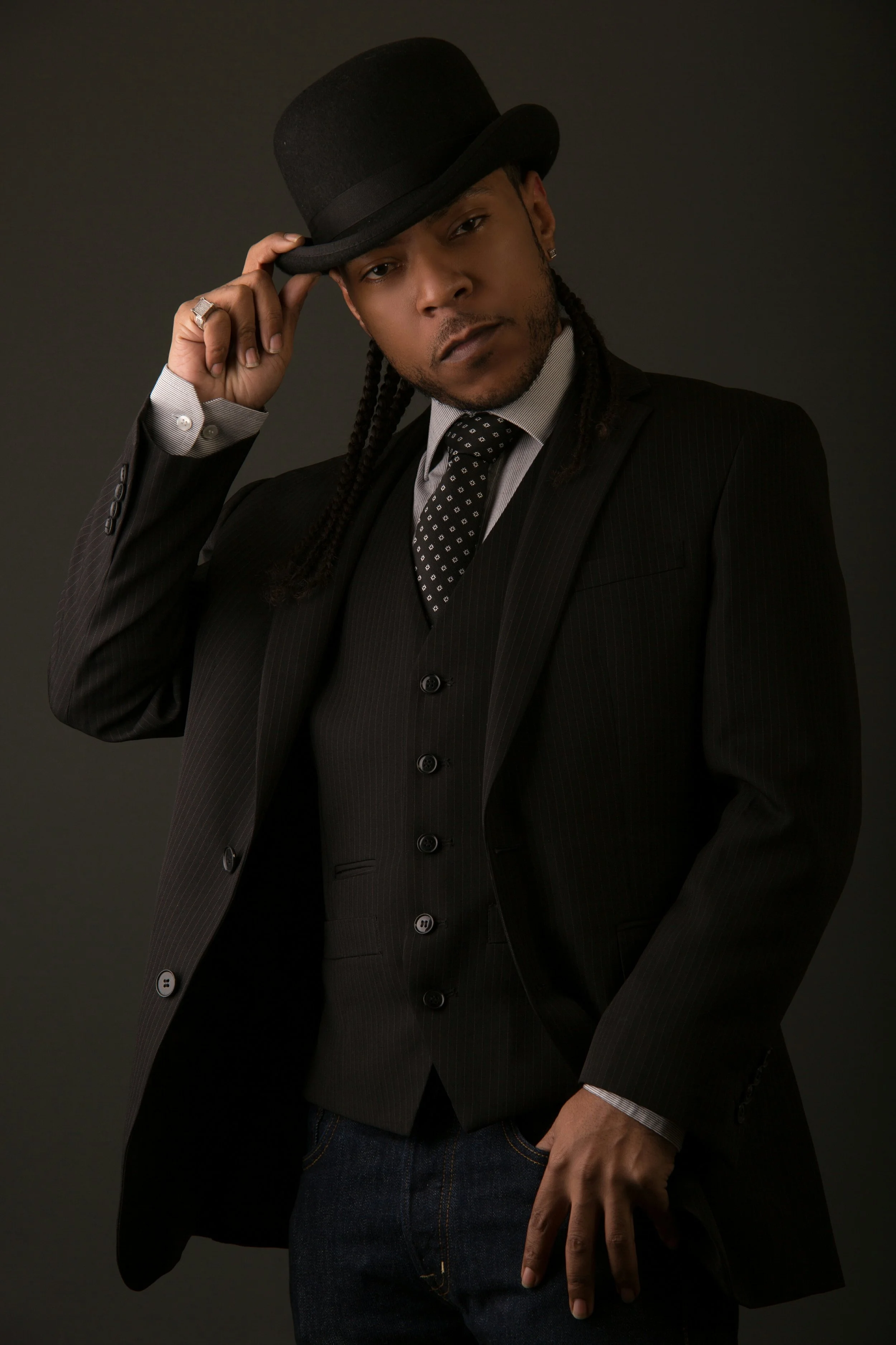 A man in a dark pinstripe suit, white shirt, and black tie with white dots, wearing a black fedora, posing against a dark background.