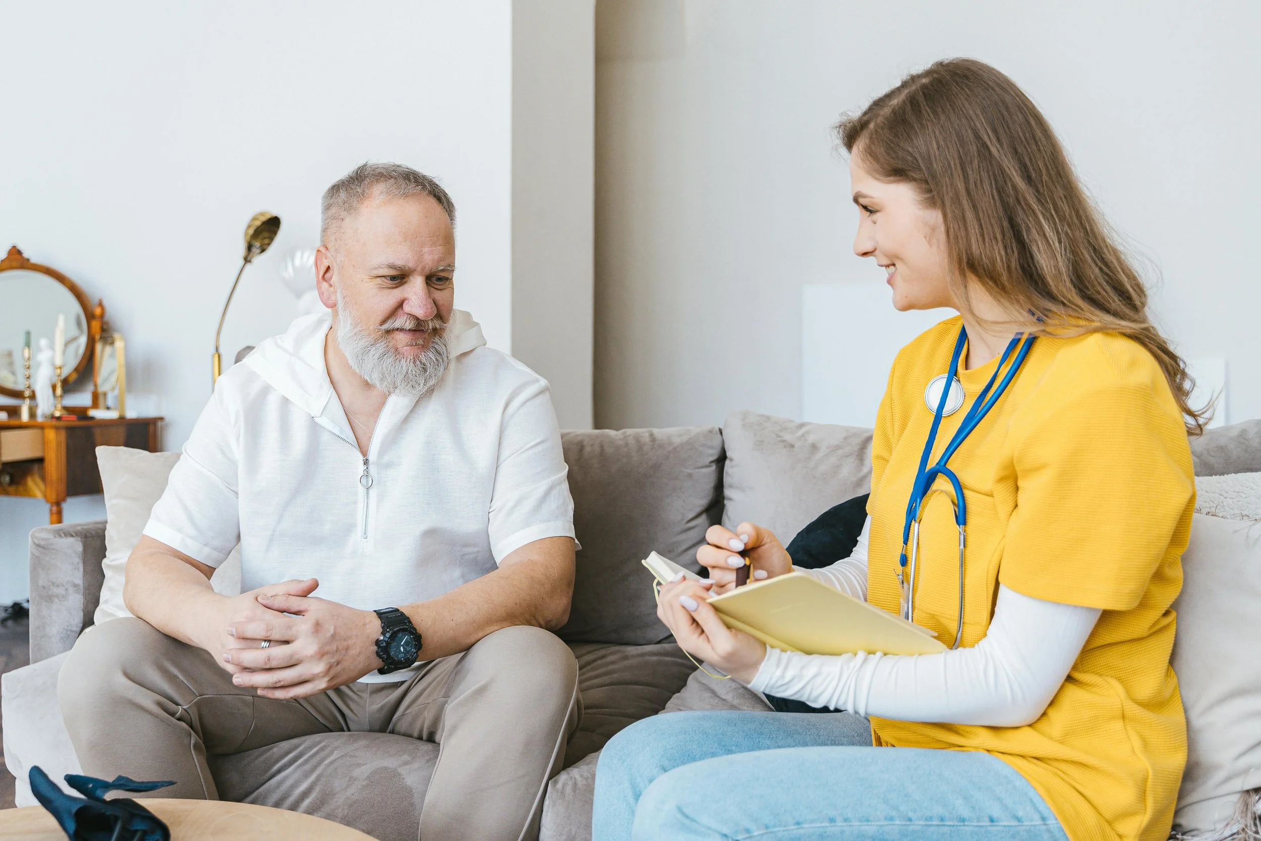 A young female nurse or healthcare professional in yellow scrubs with a stethoscope around her neck, taking notes in a yellow notepad, talking to an older man with a white beard and light hair, sitting on a sofa in a living room.