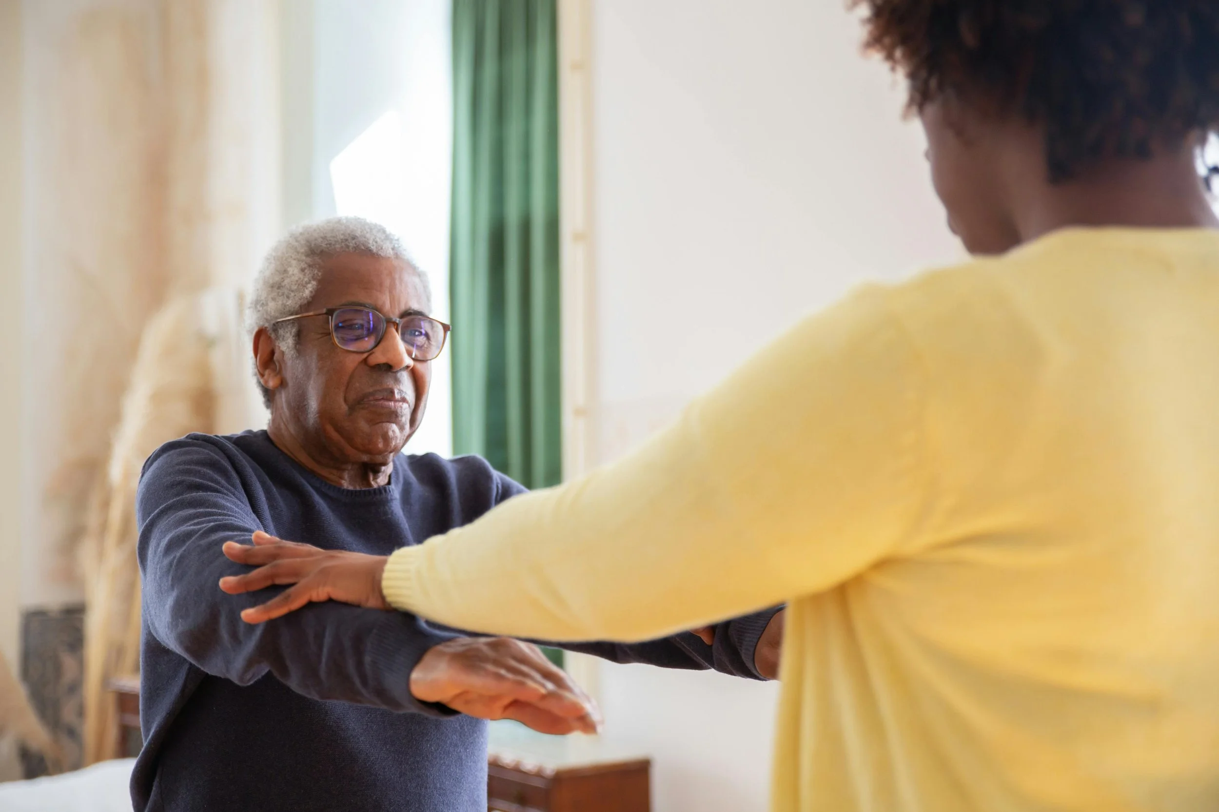 An elderly man with gray hair and glasses standing with his arms crossed, appearing cautious or hesitant, as a woman in a yellow sweater reaches out to him in a room with natural light and green curtains.