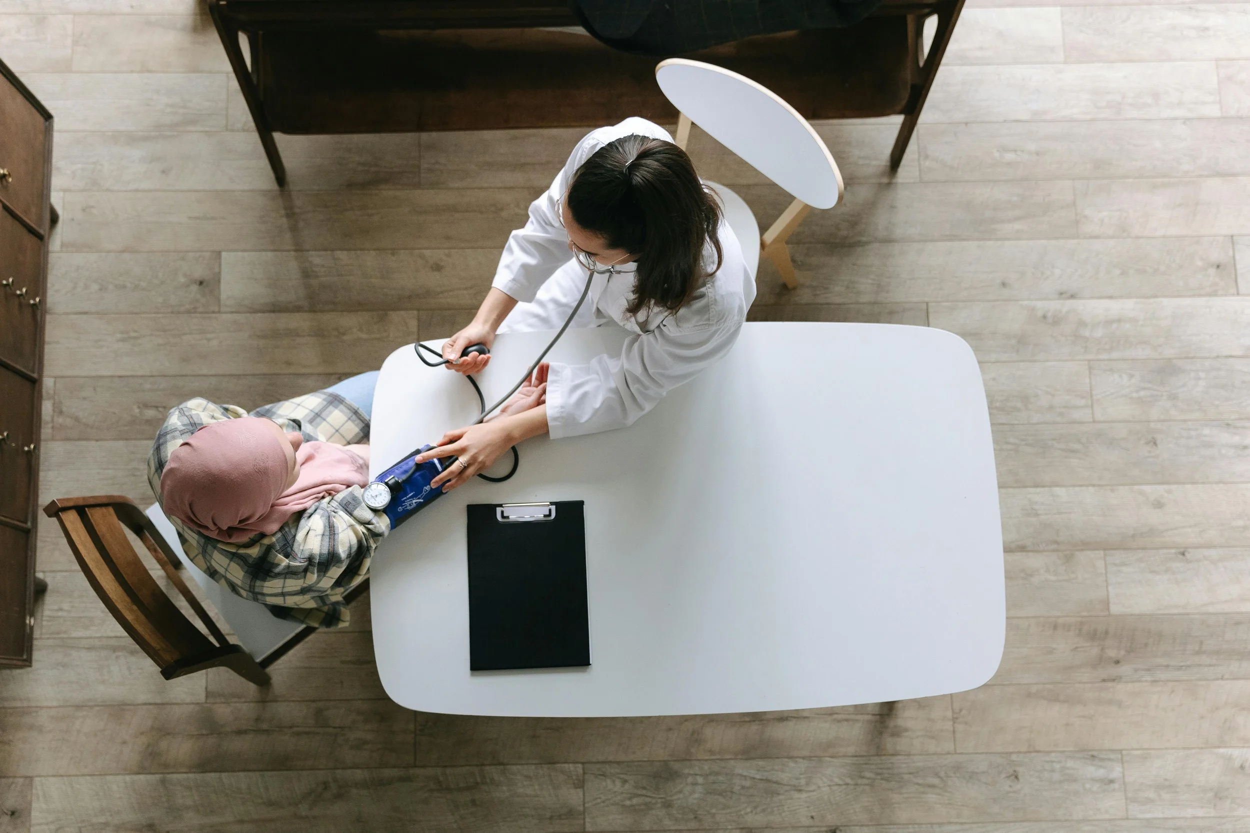 A healthcare professional measures a patient's blood pressure in a medical setting, with the healthcare worker wearing a white coat and the patient dressed in a plaid shirt and headscarf.