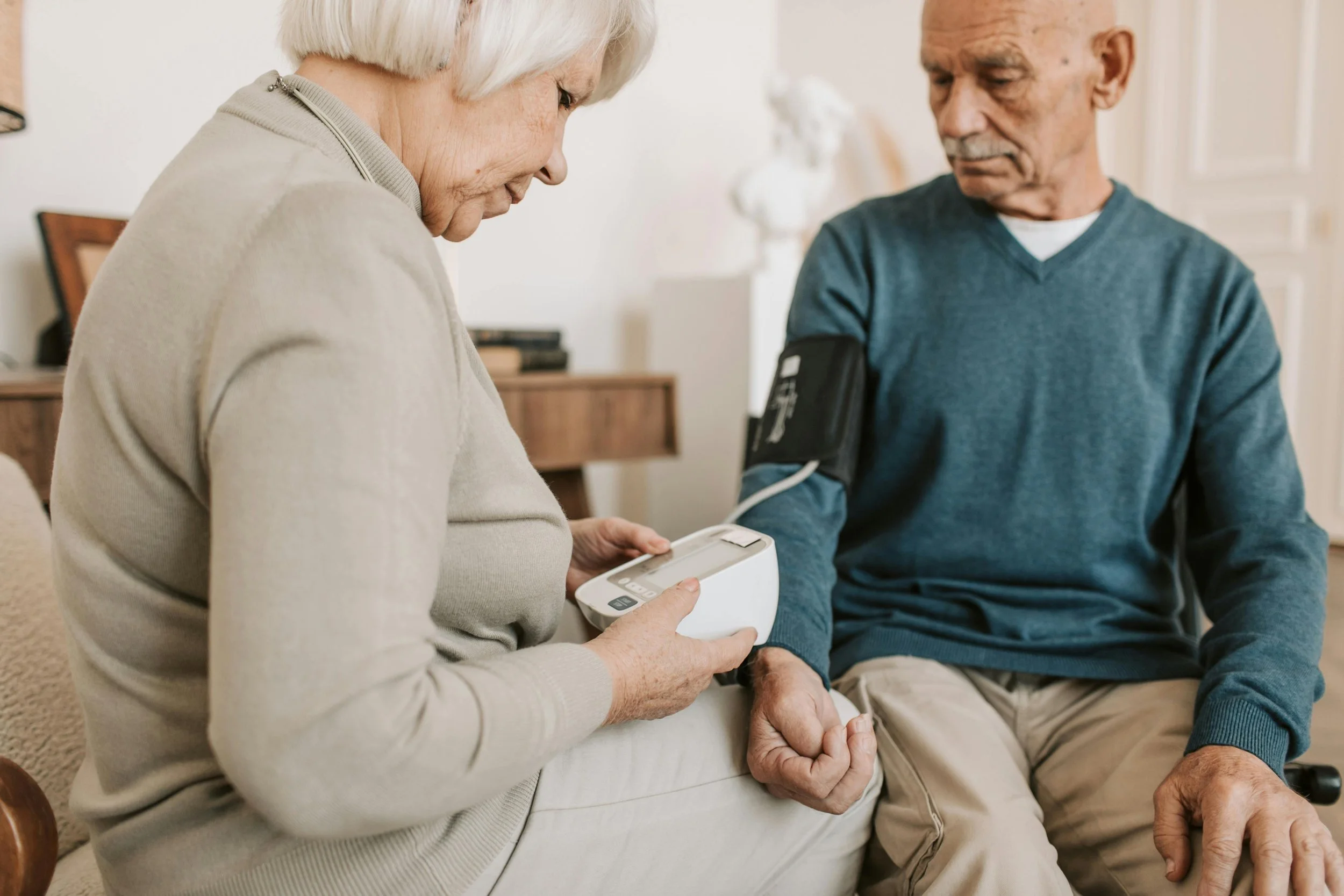 An elderly woman measures blood pressure of an elderly man using a digital blood pressure monitor, sitting on a sofa in a home setting.