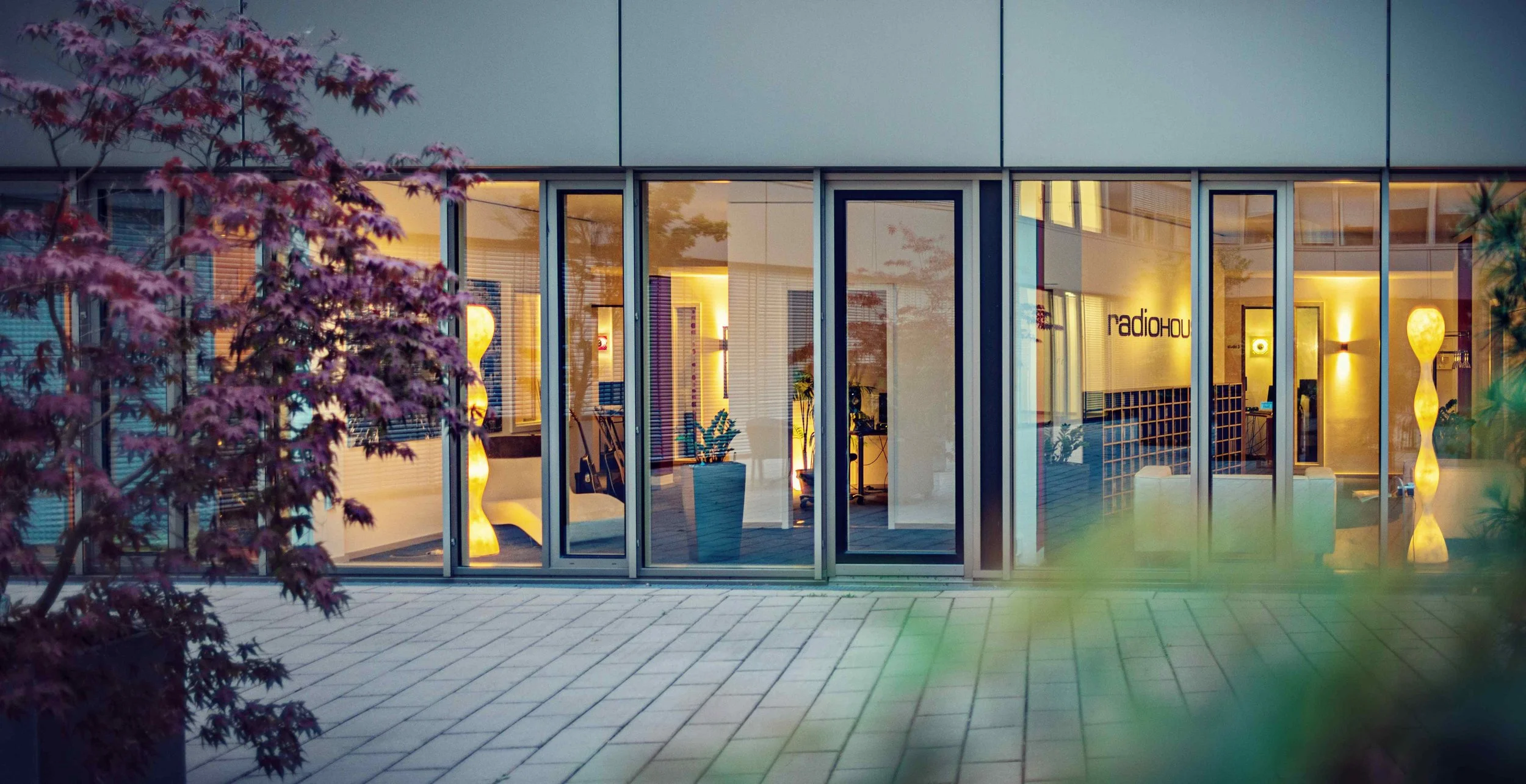 Blick durch ein Glastür- und Fensterfront auf ein modern eingerichtetes Office mit Stehlampen, Pflanzen und dem Schriftzug 'radiohouse' an der Wand.