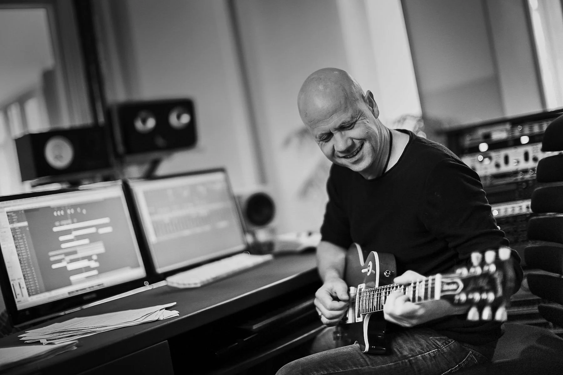 Mann spielt Gitarre in einem Tonstudio, umgeben von Computerbildschirmen und Tontechnik.