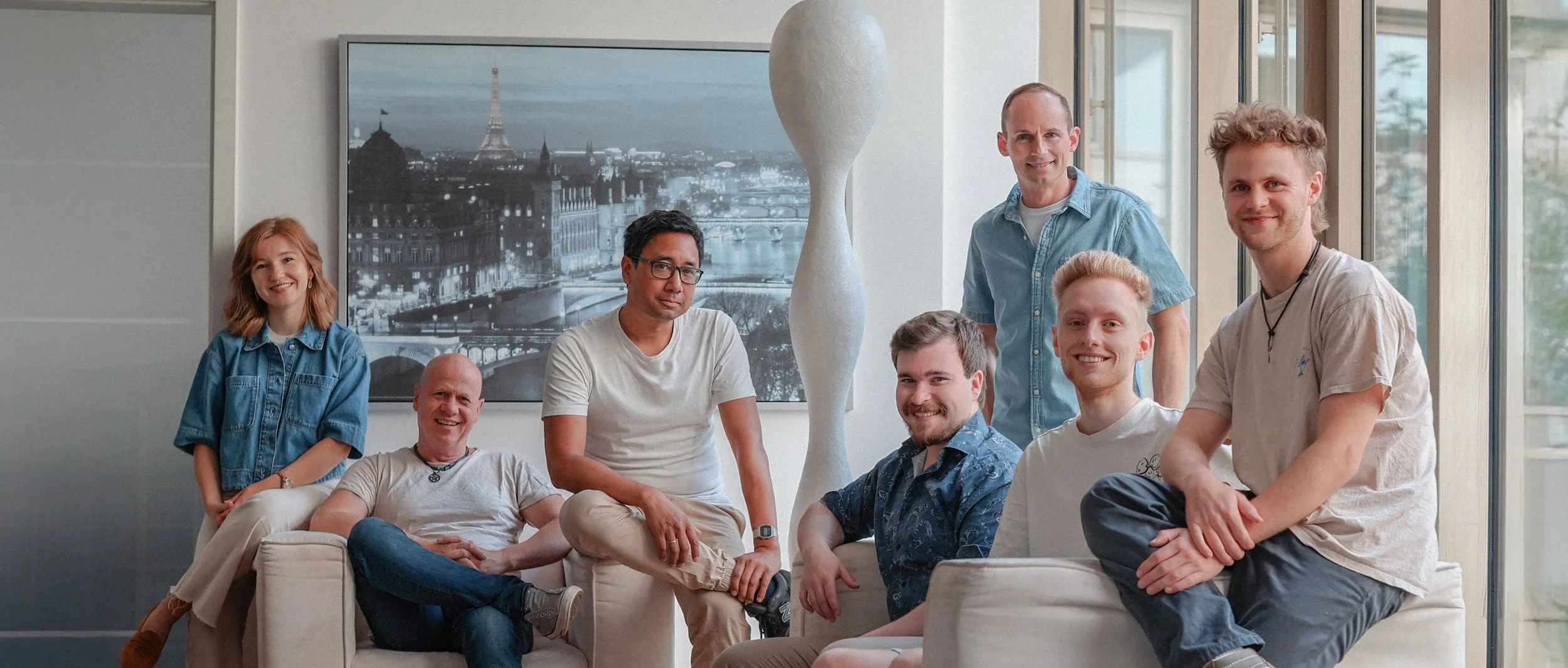 Eine Gruppe von sieben jungen Erwachsenen, sechs Männer und einer Frau, posieren fröhlich in einem modernen, gut beleuchteten Raum mit großen Fenstern und einer Pariser Stadtlandschaft im Hintergrund.
