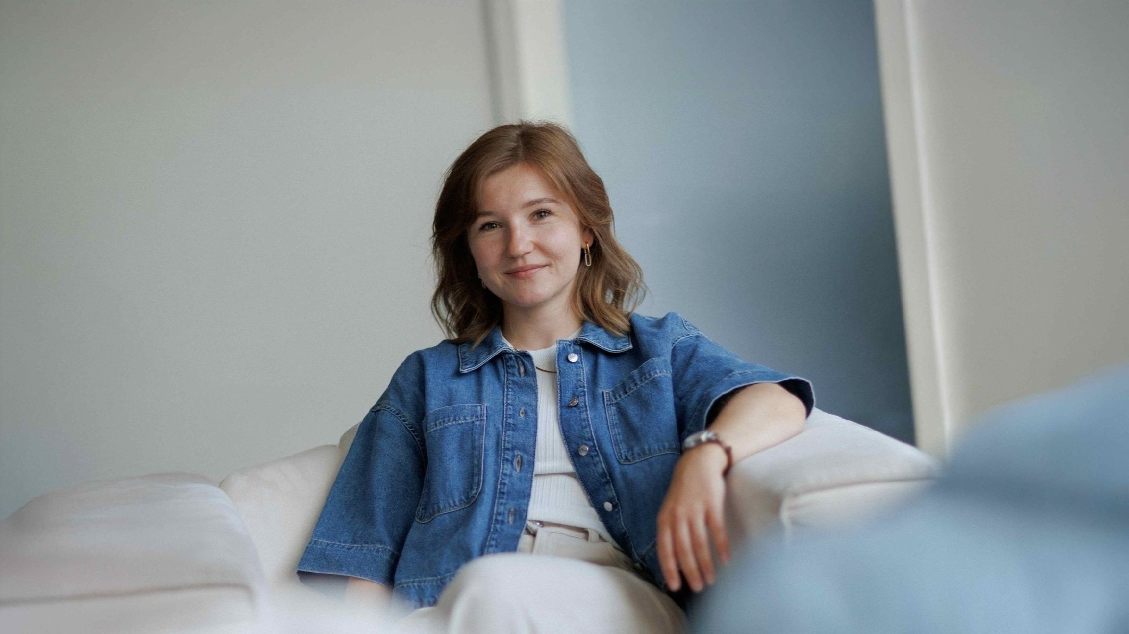 Junge Frau mit braunen Haaren lächelnd auf einem weißen Sofa sitzend, trägt eine blaue Denim-Jacke und weiße Hose.