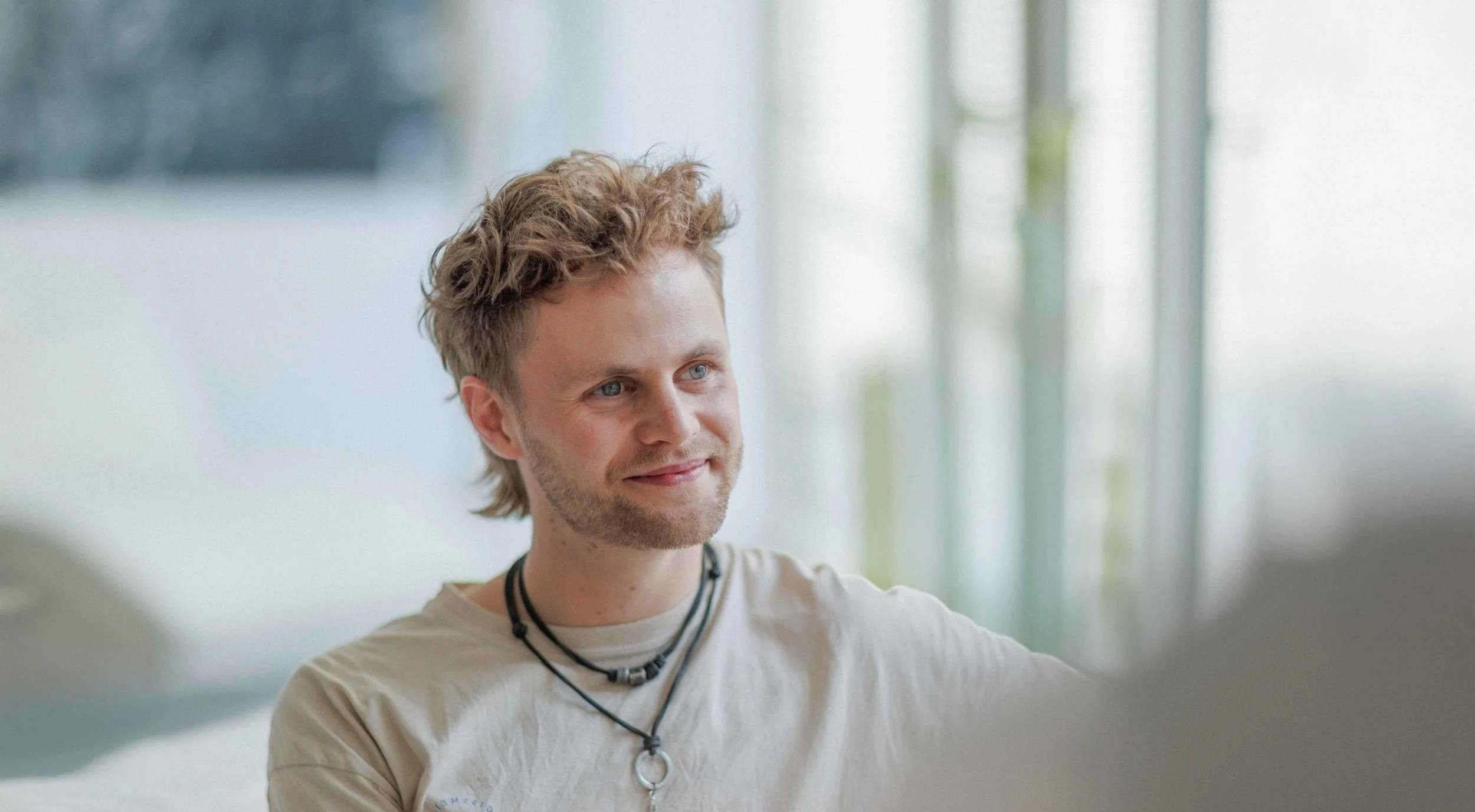 Ein junger Mann mit blondem, leicht lockigem Haar lächelt und sitzt in einem hellen Raum, möglicherweise im Gespräch.