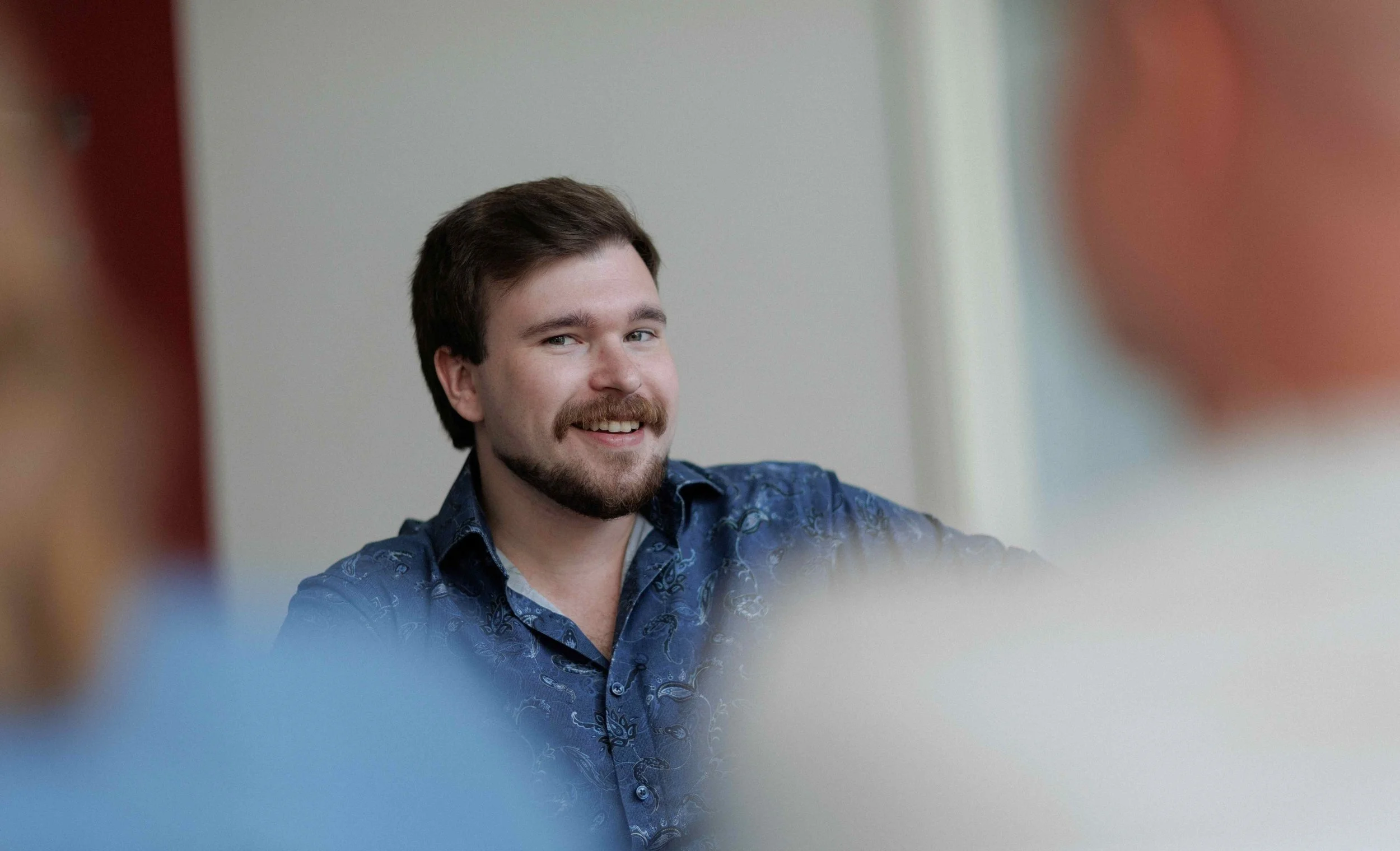 Ein lachender Mann mit dunklem Haar und Bart, trägt ein blaugemustertes Hemd, sitzt in einem Raum.