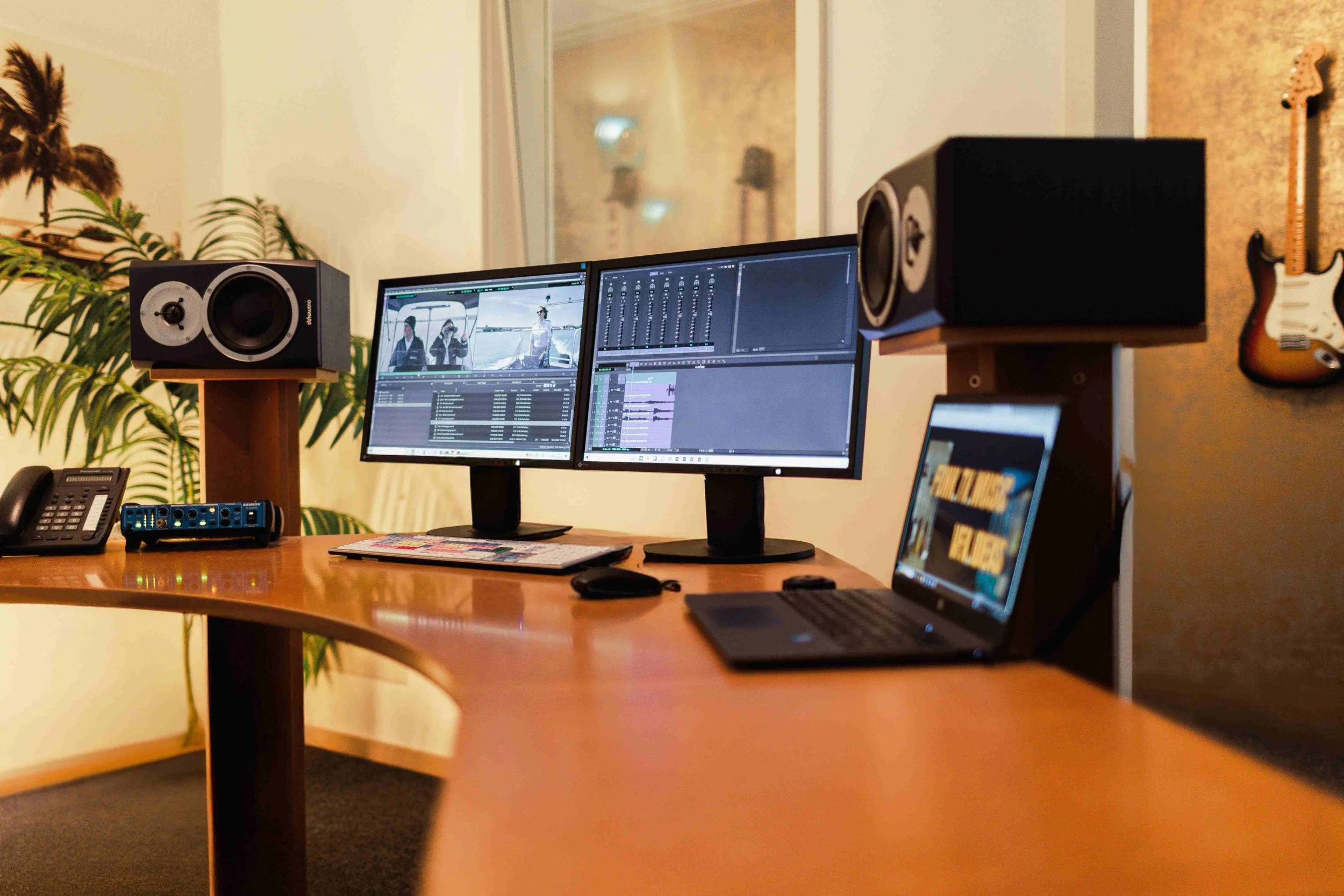 Arbeitsplatz mit doppelt monitore, Lautsprechern, Laptop, Telefon, Pflanzen, Gitarre an der Wand, und einem Mischpult.