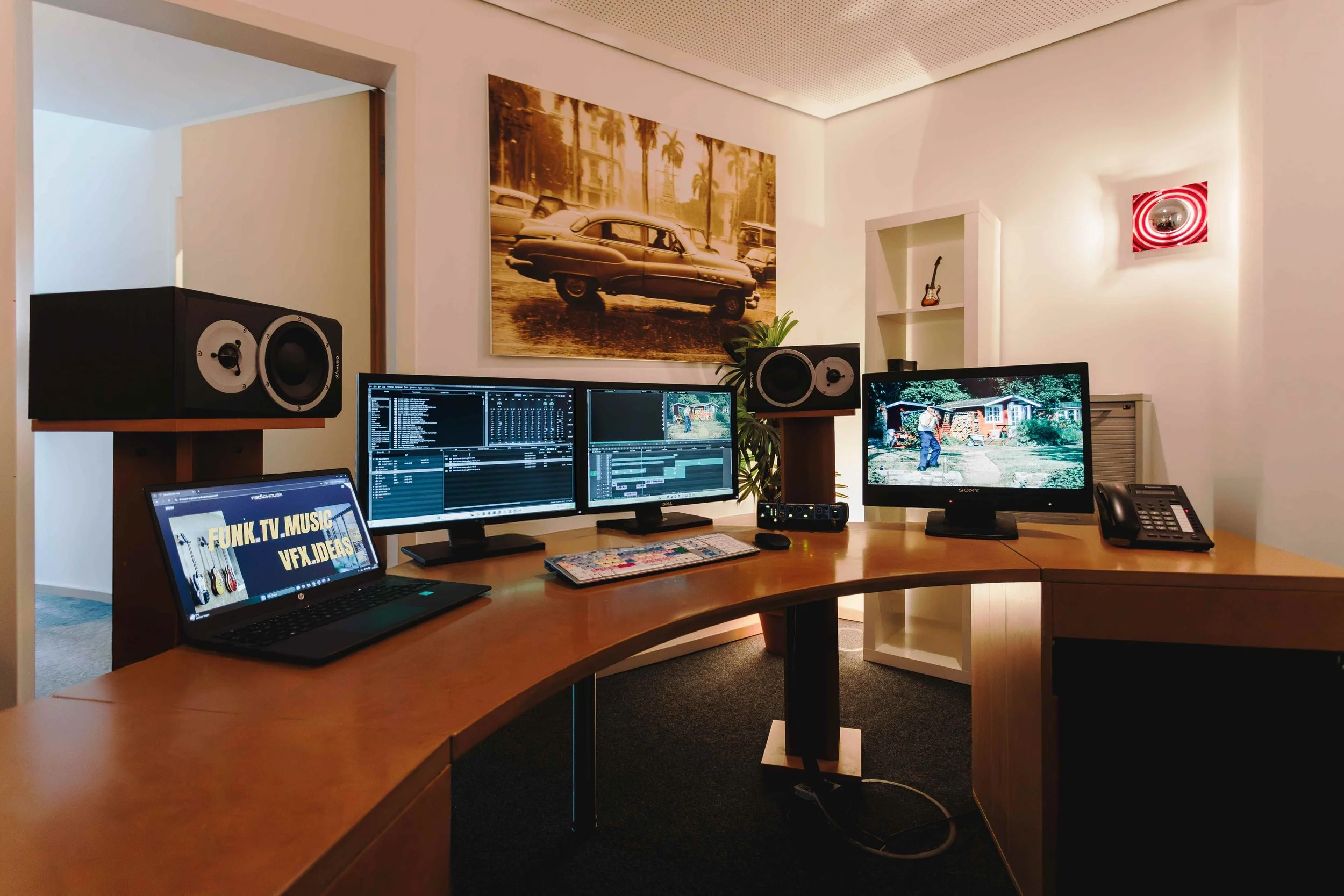 Arbeitsplatz mit mehreren Monitoren, einem Laptop, professionellen Lautsprechern und Bürotelefon in einem Raum mit Wanddekorationen, einem Kunstwerk eines Autos, und einem kleinen Regal.