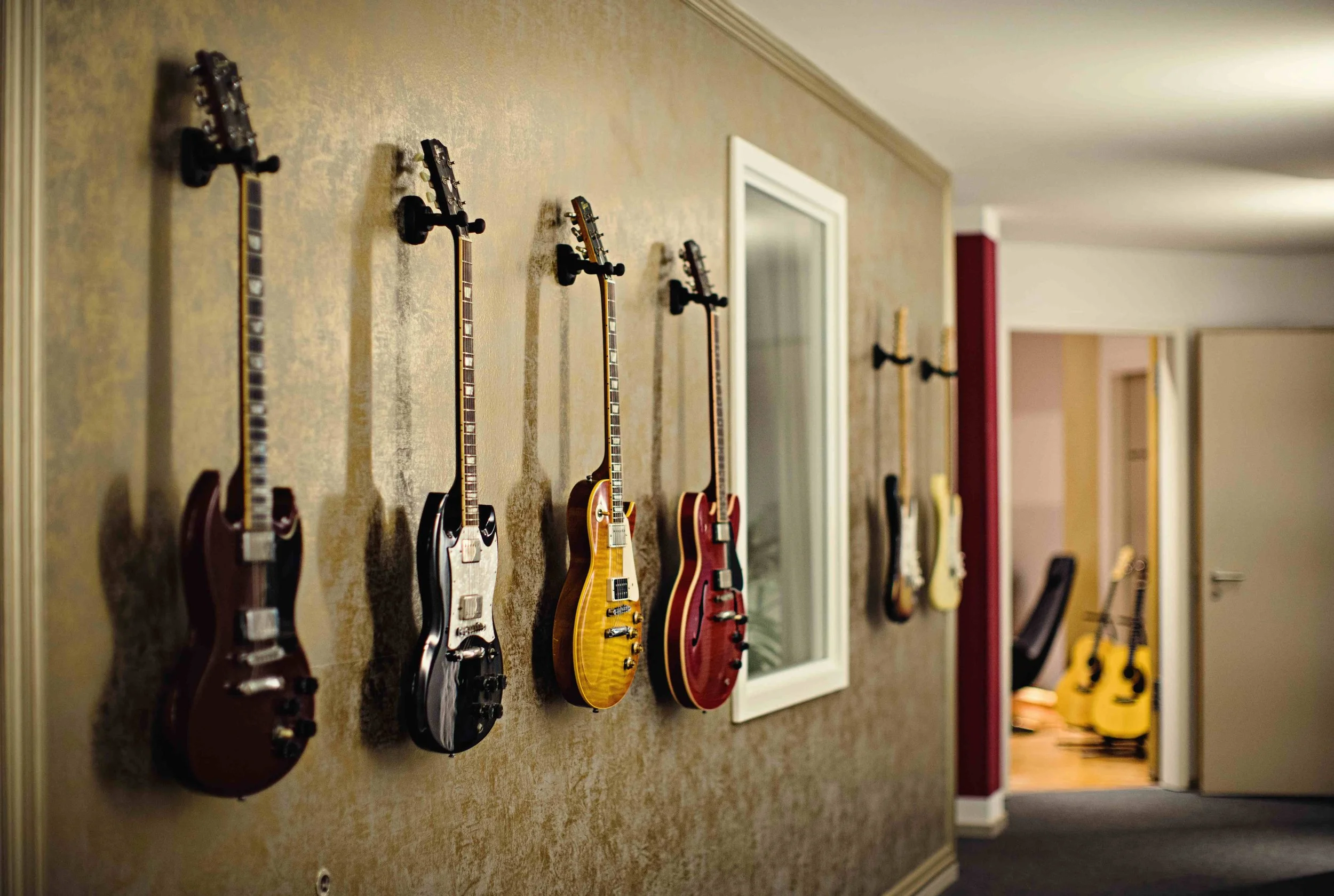 Mehrere E-Gitarren hängen an einer Wand in einem Musikraum, im Hintergrund weitere Gitarren stehen auf dem Boden.