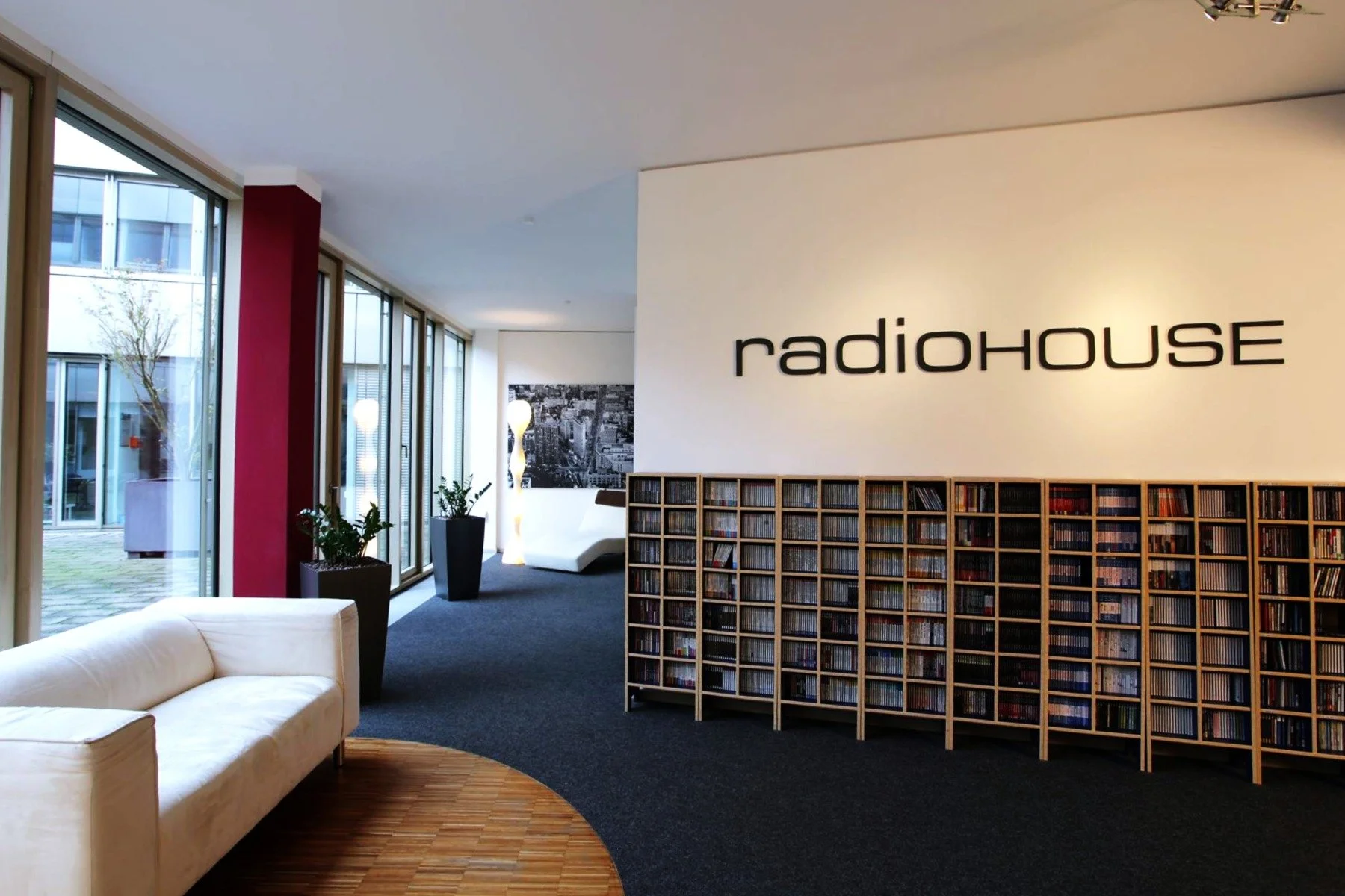 Innenraum eines Gebäudes mit Sitzbereich, Bücherregalen, Pflanzen und großer Beschriftung 'radioHOUSE' an der Wand.