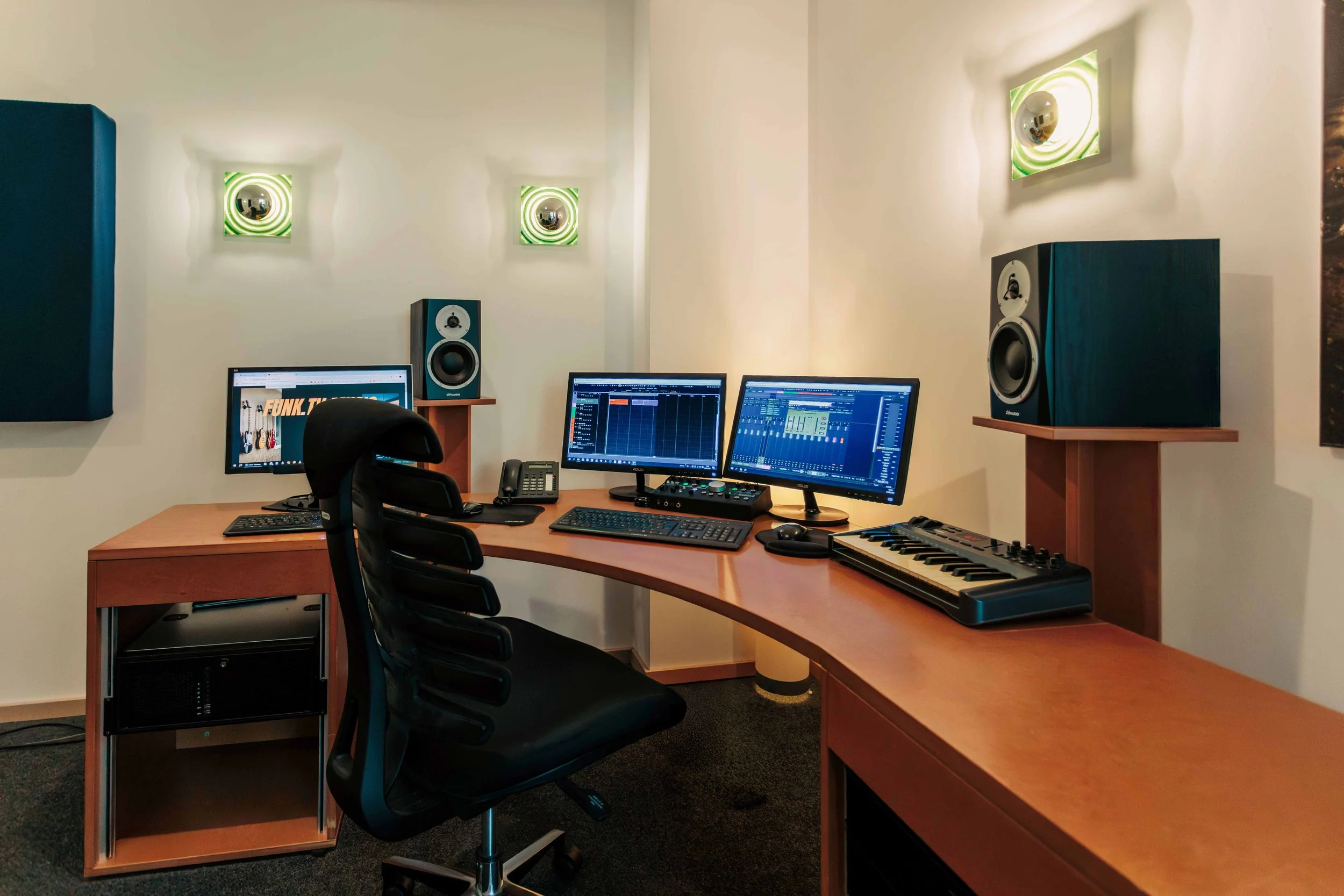 Aufnahme eines Musikproduktionsraums mit drei Monitore, Studio-Lautsprechern, Musik-Software auf den Bildschirmen, einem Keyboard, einem Mikrofon, einem Telefon und einem Computer.