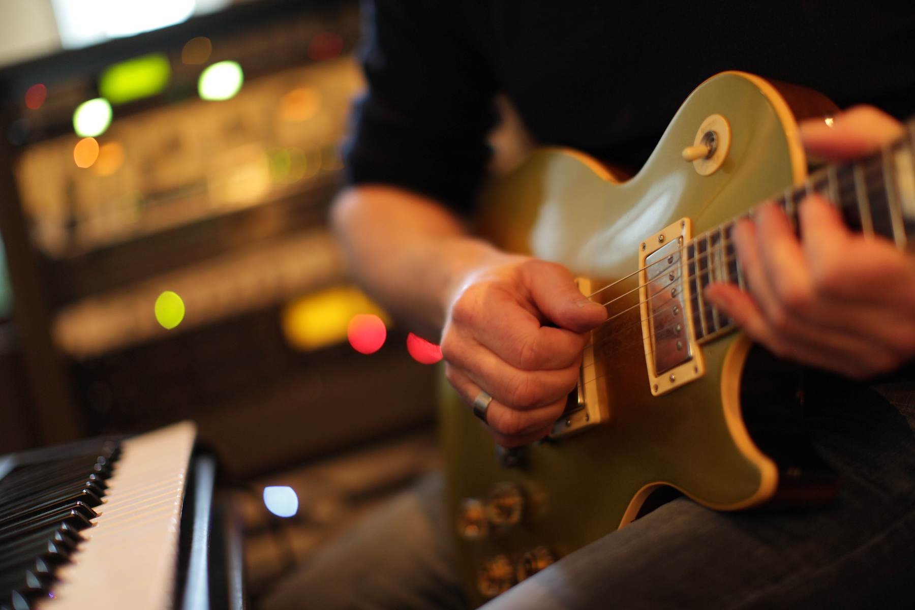 Person spielt E-Gitarre in einem Musikstudio, mit einem Keyboard im Vordergrund und bunten Lichter im Hintergrund.