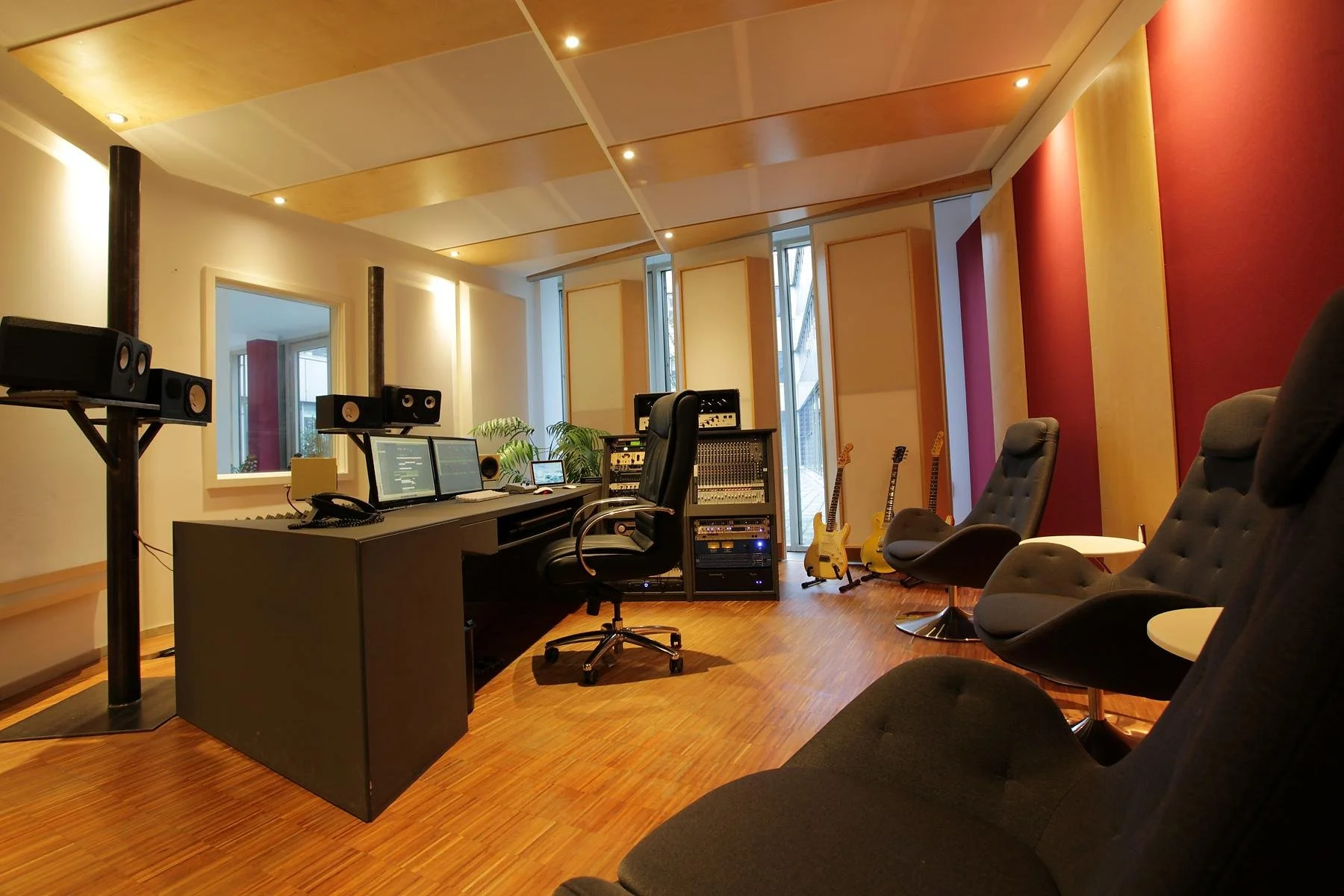 Aufnahme eines Tonstudios mit Schreibtisch, mehreren Monitoren, einem Chefsessel, akustischen Panels an den Wänden, Gitarre- und Bassinstrumenten, sowie mehrere Lounge-Stühle.