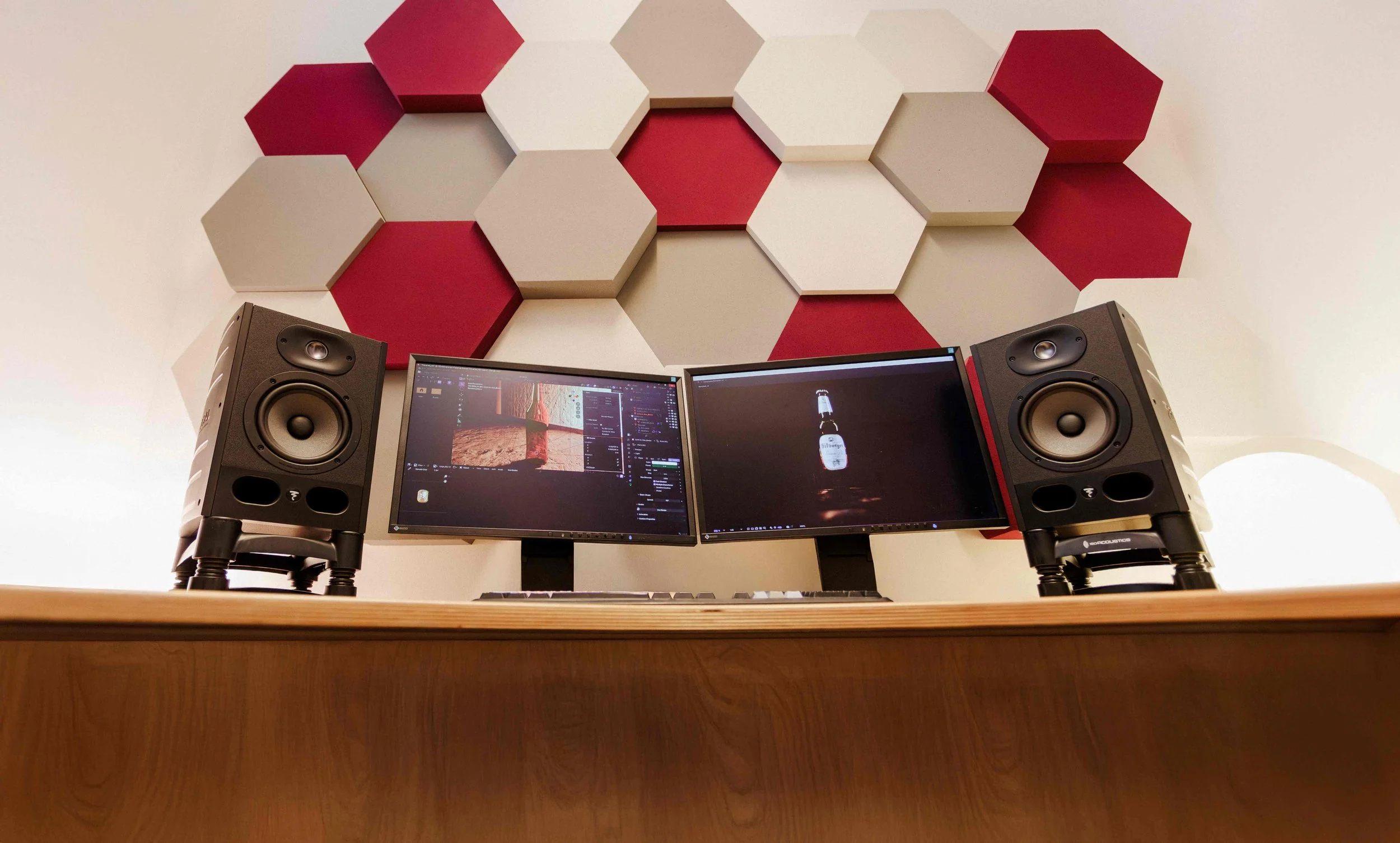 Zwei Lautsprecher, zwei Monitore mit Bildschirmschoner, Tastatur auf einem Holzschreibtisch, hinter einem Wandpaneel mit roten und weißen Hexagon-Formen.