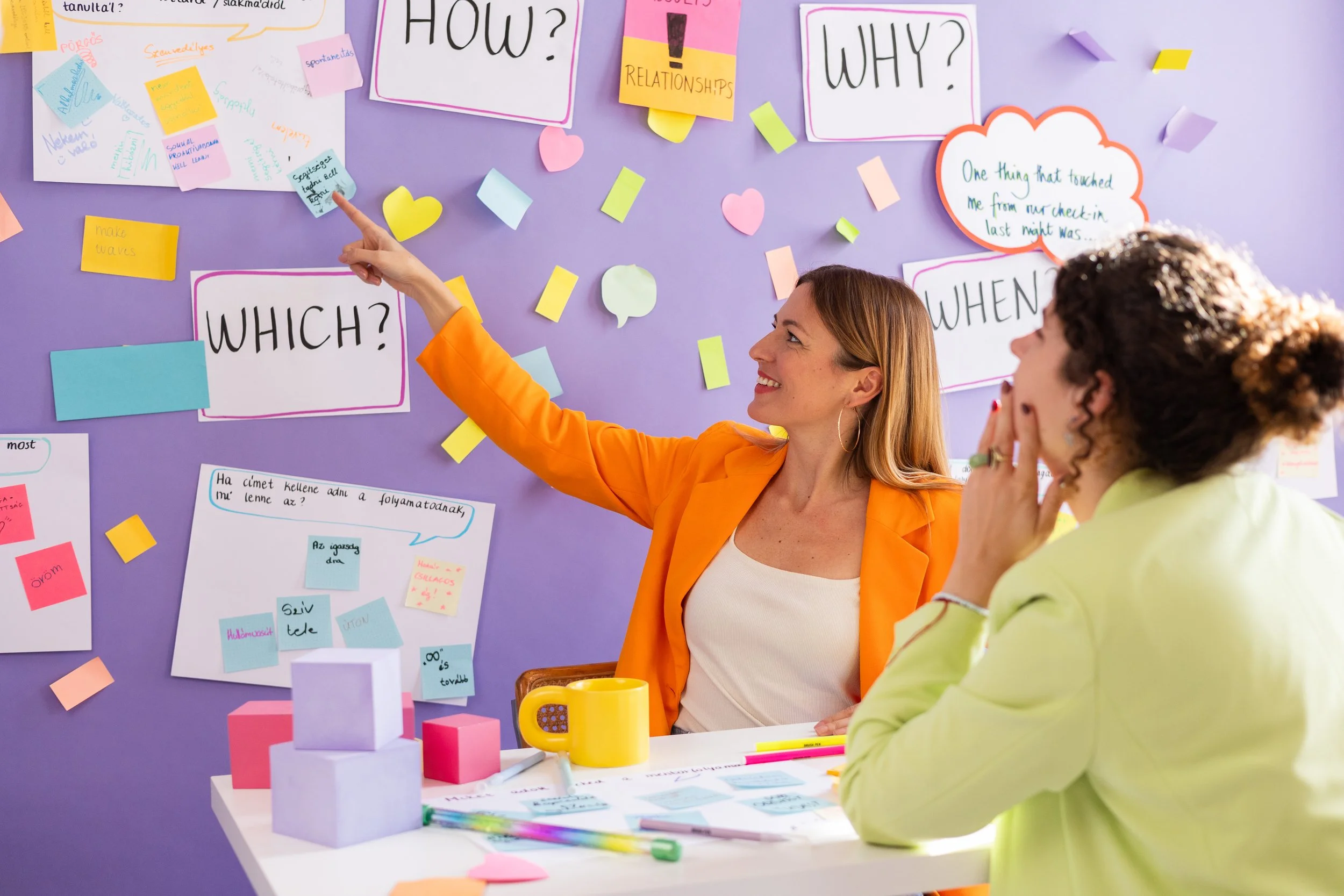 Two women are engaged in a discussion in front of a purple wall covered with colorful sticky notes, signs, and handwritten notes about questions like "HOW?", "WHY?", "WHICH?", and "WHEN?". One woman, wearing an orange blazer, is smiling and pointing at the wall, while the other woman, with curly hair and a light green shirt, listens attentively. There are various stationery and small objects on a white table in front of them.