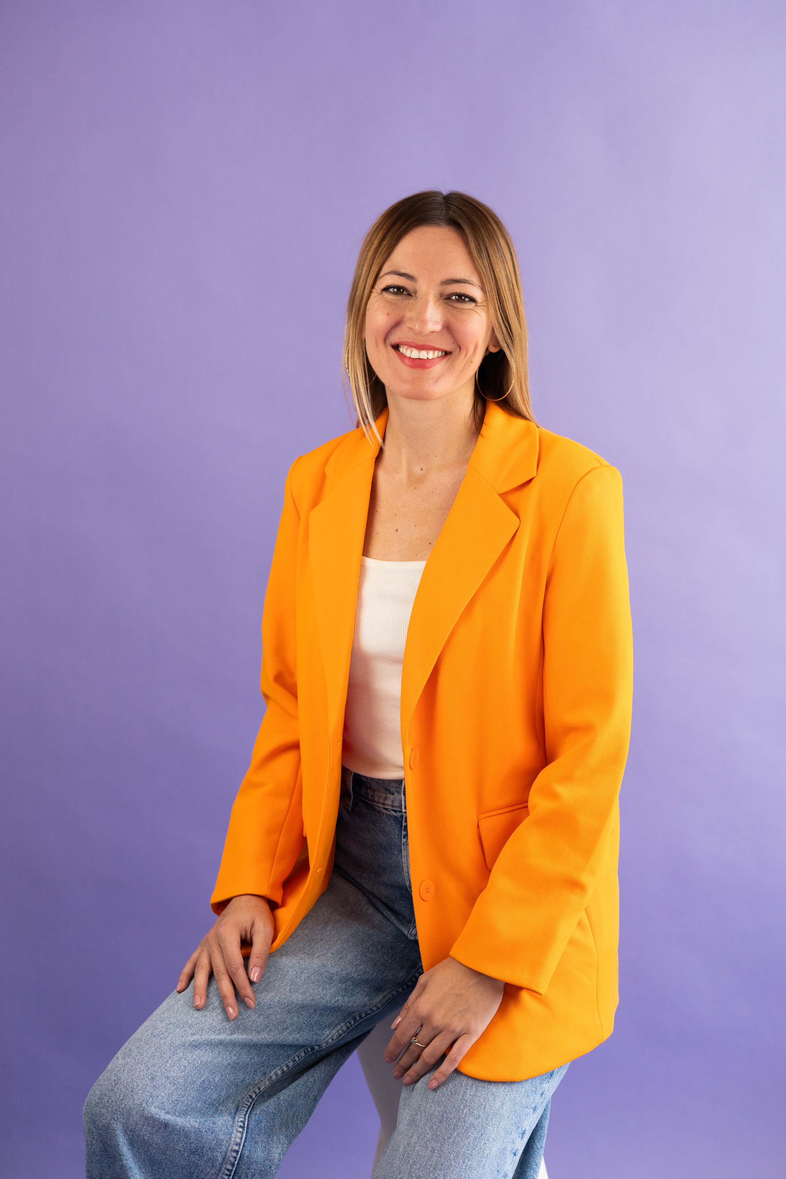 A woman with shoulder-length hair and hoop earrings, smiling, sitting against a purple background, wearing a bright yellow blazer over a white top, with light blue jeans.
