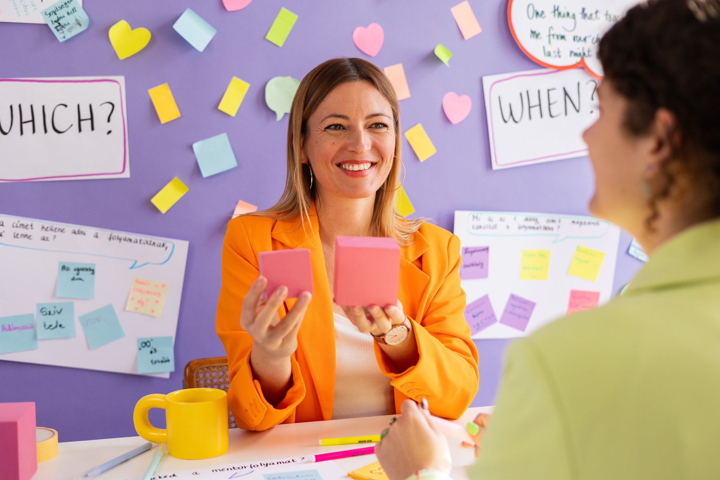 A woman with brown hair, wearing an orange blazer and earrings, is smiling and holding pink sticky notes. She is sitting at a table with yellow and pink mugs, pens, and paper. Behind her, there is a purple wall decorated with colorful paper notes, hearts, and speech bubbles, and visible text including questions like 'WHICH?' and 'WHEN?'.