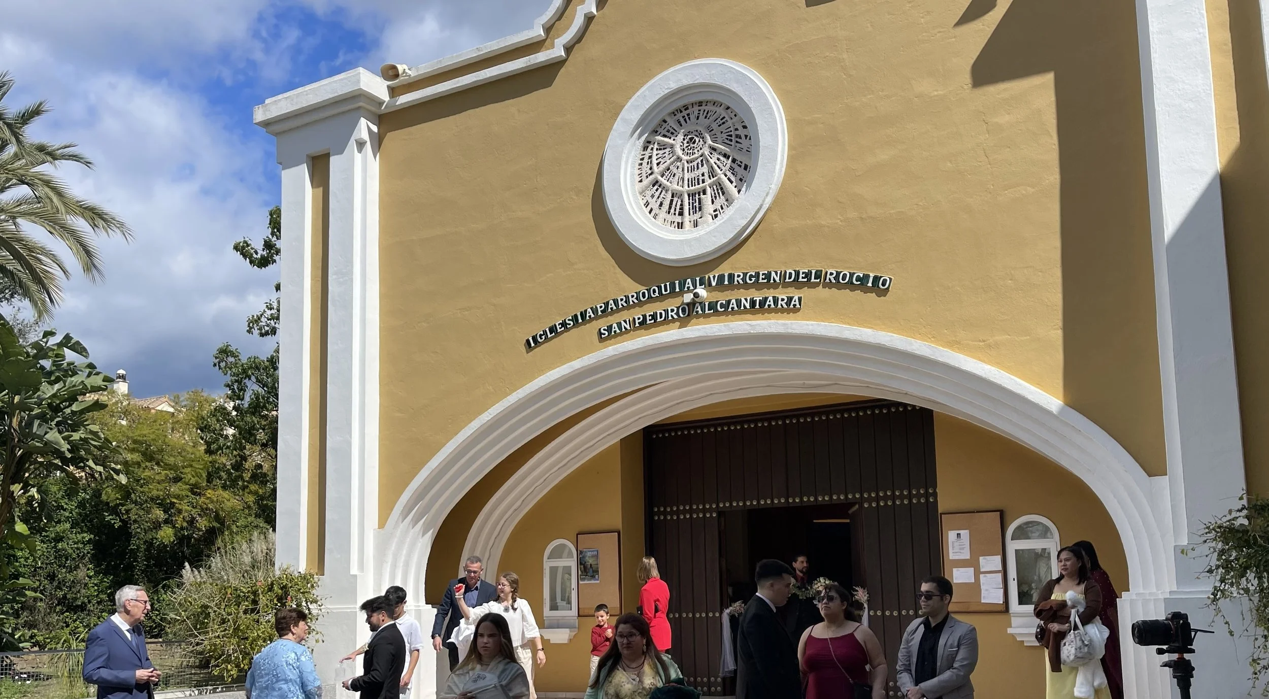 Group of people gathered outside a yellow church with white trim, arched entrance, and a round stained glass window above the door.