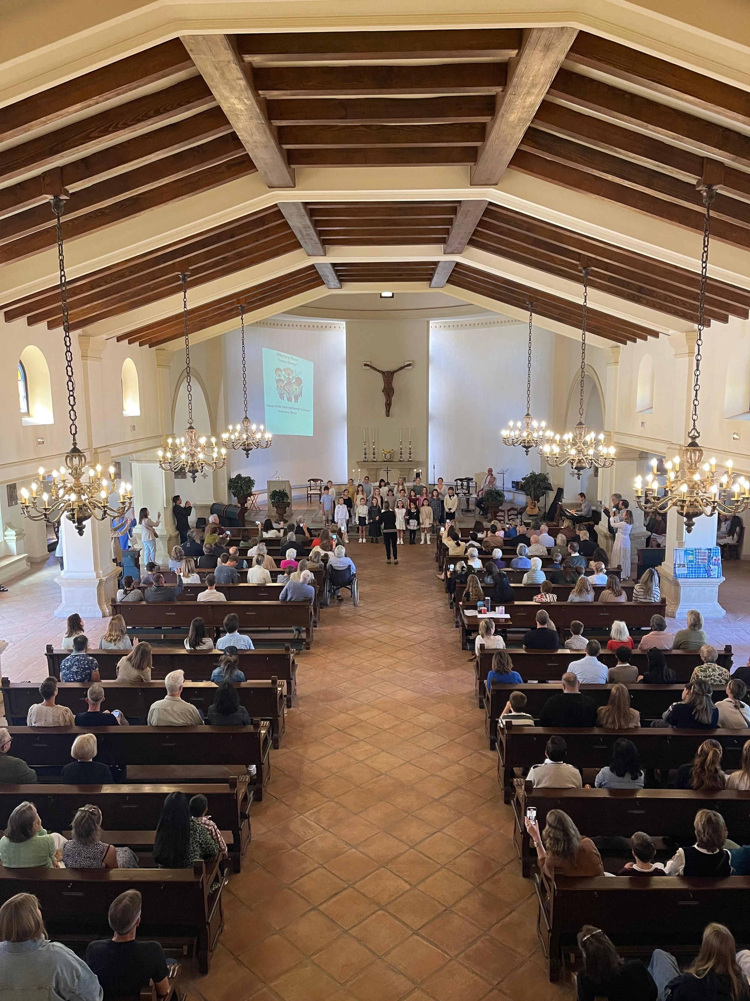 A church interior during a community event with people seated in pews and children performing at the front, decorated with chandeliers and religious symbols.