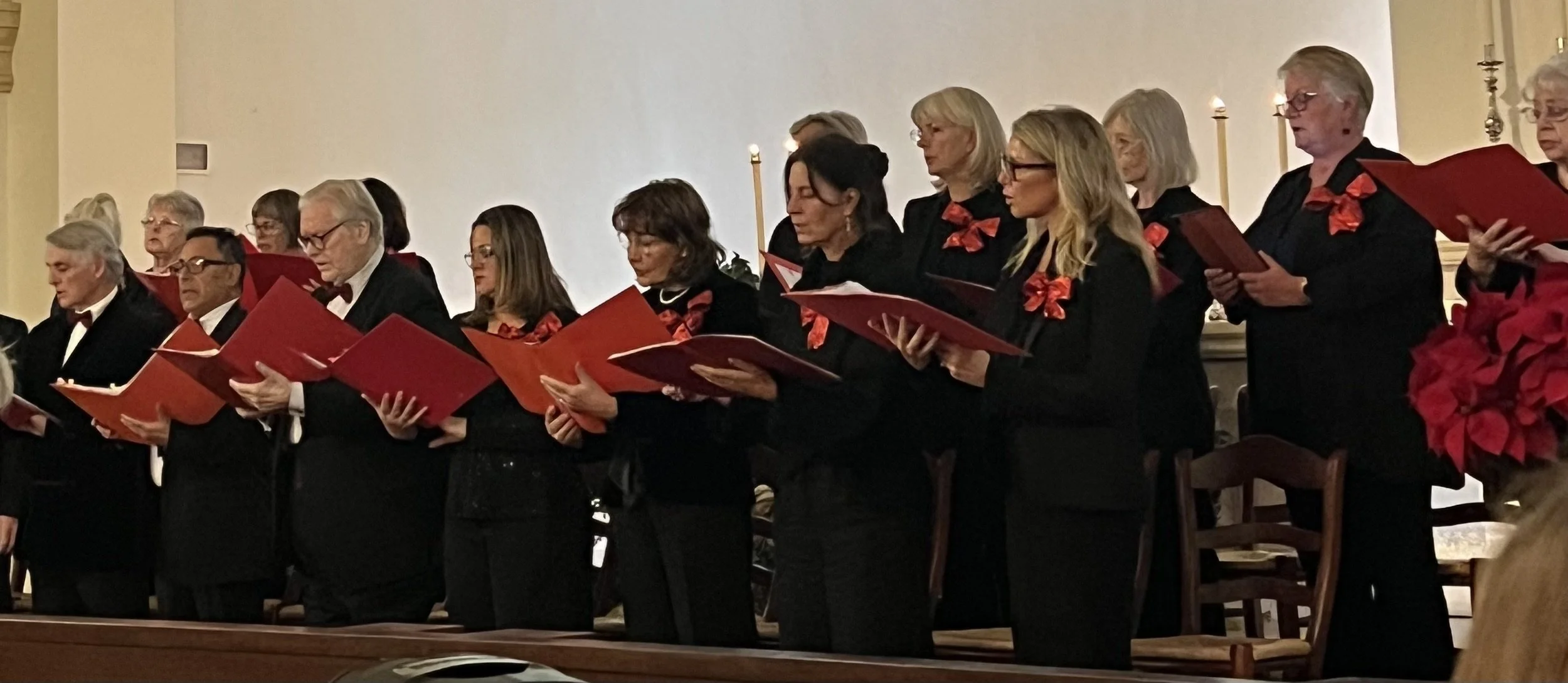 Group of elderly people standing and singing during a choir performance, holding red songbooks, in a church decorated with candles and poinsettias.