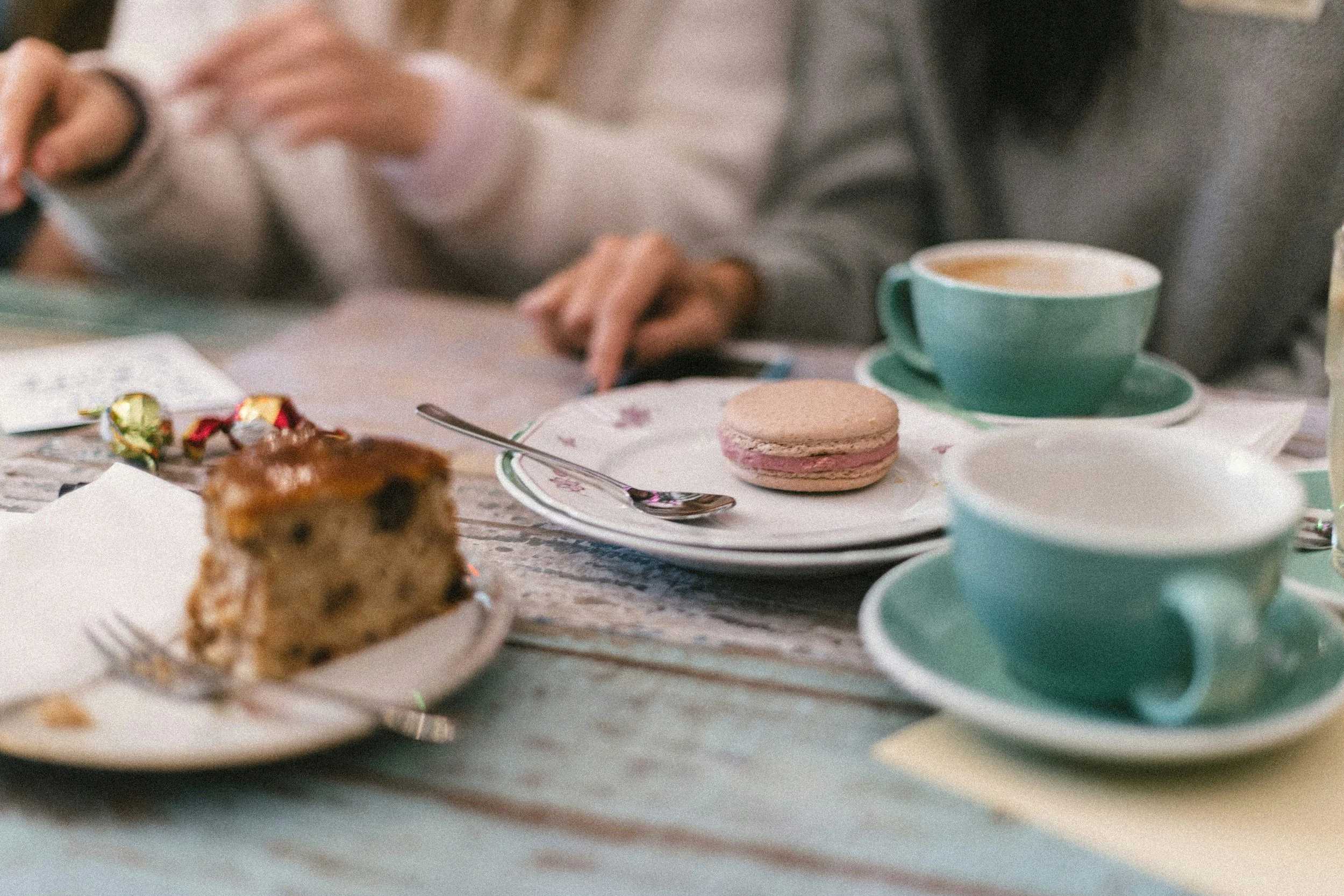 A plate with a pink macaroon, two teal cups with drinks, and a slice of fruitcake on a rustic wooden table, with two people sitting in the background.