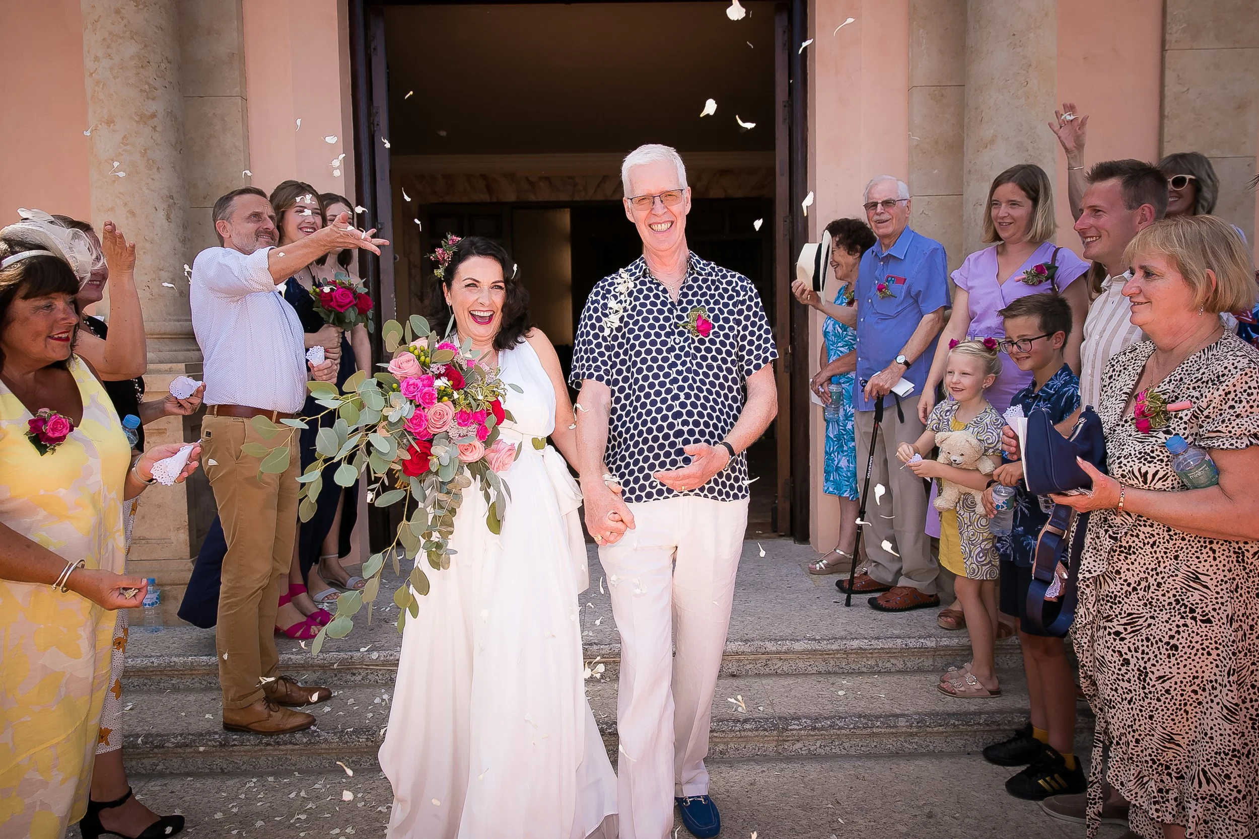 Wedding couple holding hands and smiling as confetti falls, surrounded by friends and family outside a building.