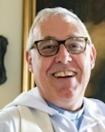 A smiling older man with white hair, glasses, and light skin, wearing a white clergy or religious robe, standing indoors against a room with paintings and decorations.