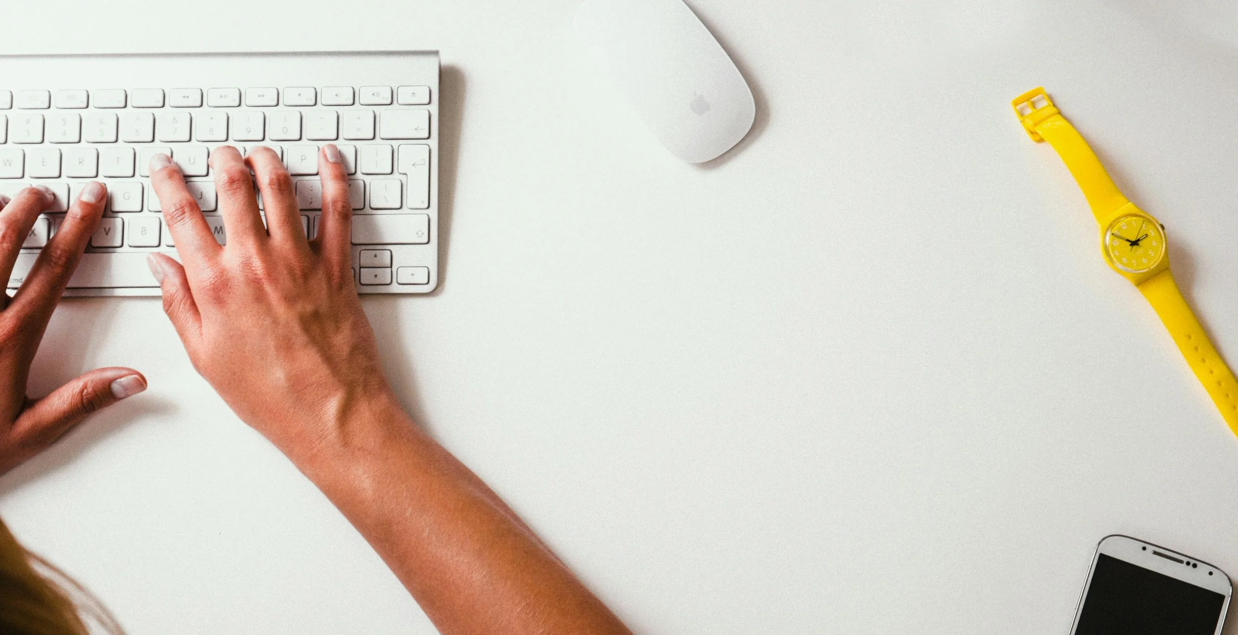 Person typing on a white keyboard on a white desk, with a white Apple Magic Mouse, yellow wristwatch, and smartphone on the desk.