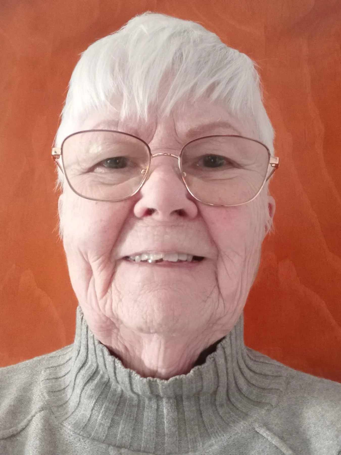 A close-up selfie of a woman with short white hair, wearing glasses and a gray turtleneck sweater, smiling in front of an orange background. This is our safeguarding officer Jean