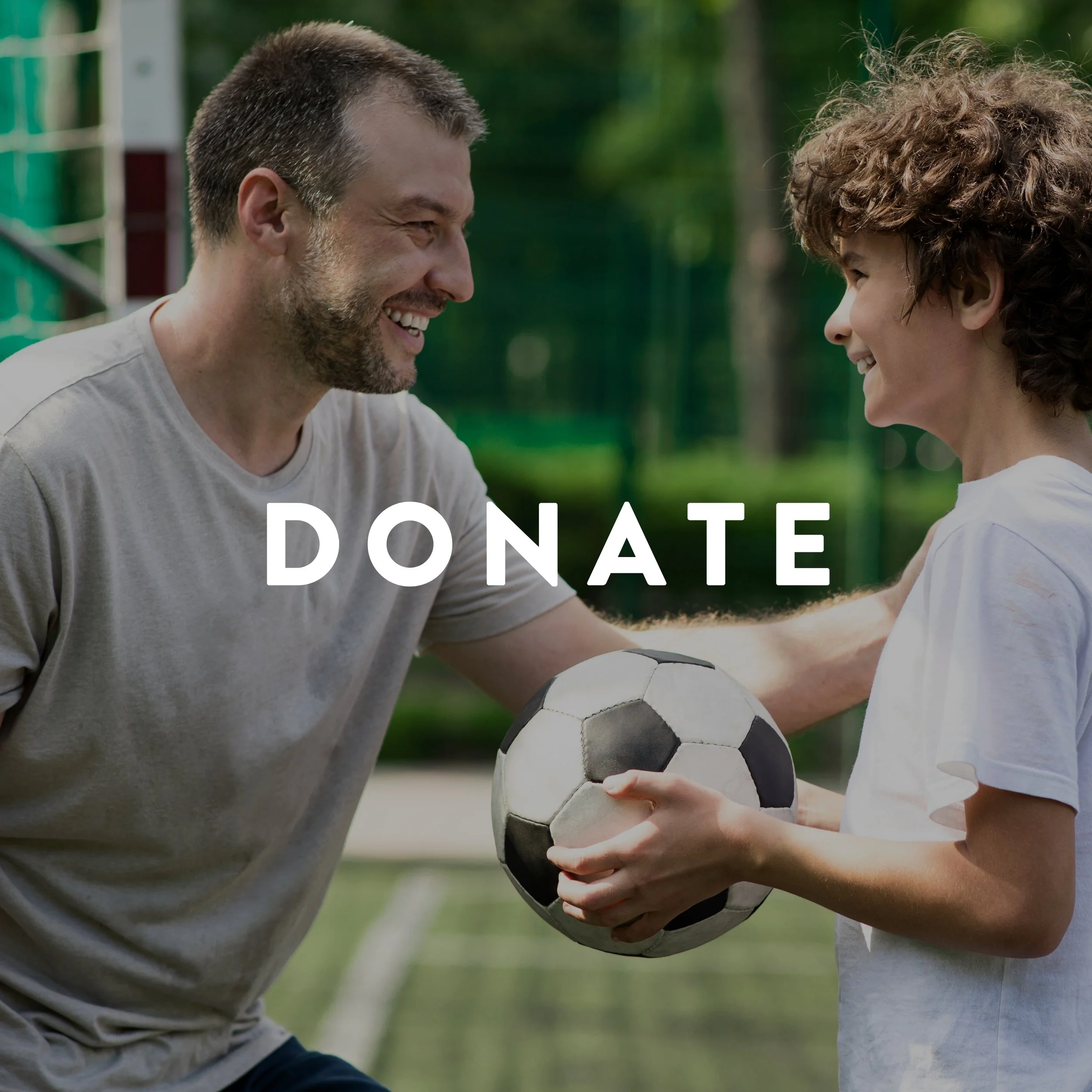 A man and a boy smiling at each other while holding a soccer ball, outdoors in a park or sports field, with the word 'DONATE' overlayed on the image.