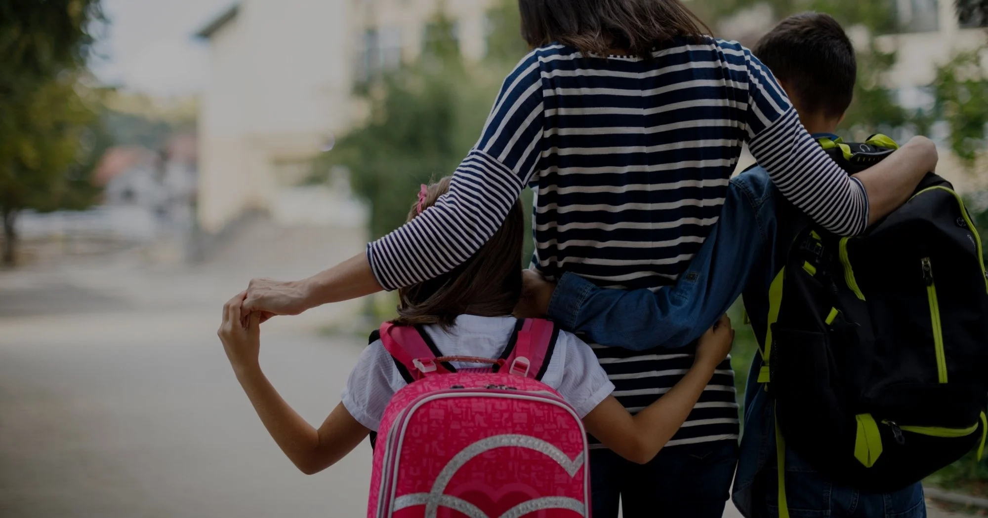 Two children, a boy and a girl, walking outdoors with backpacks, holding hands, and the girl has her arm around the boy's shoulders. The scene appears to be in a neighborhood or park setting.